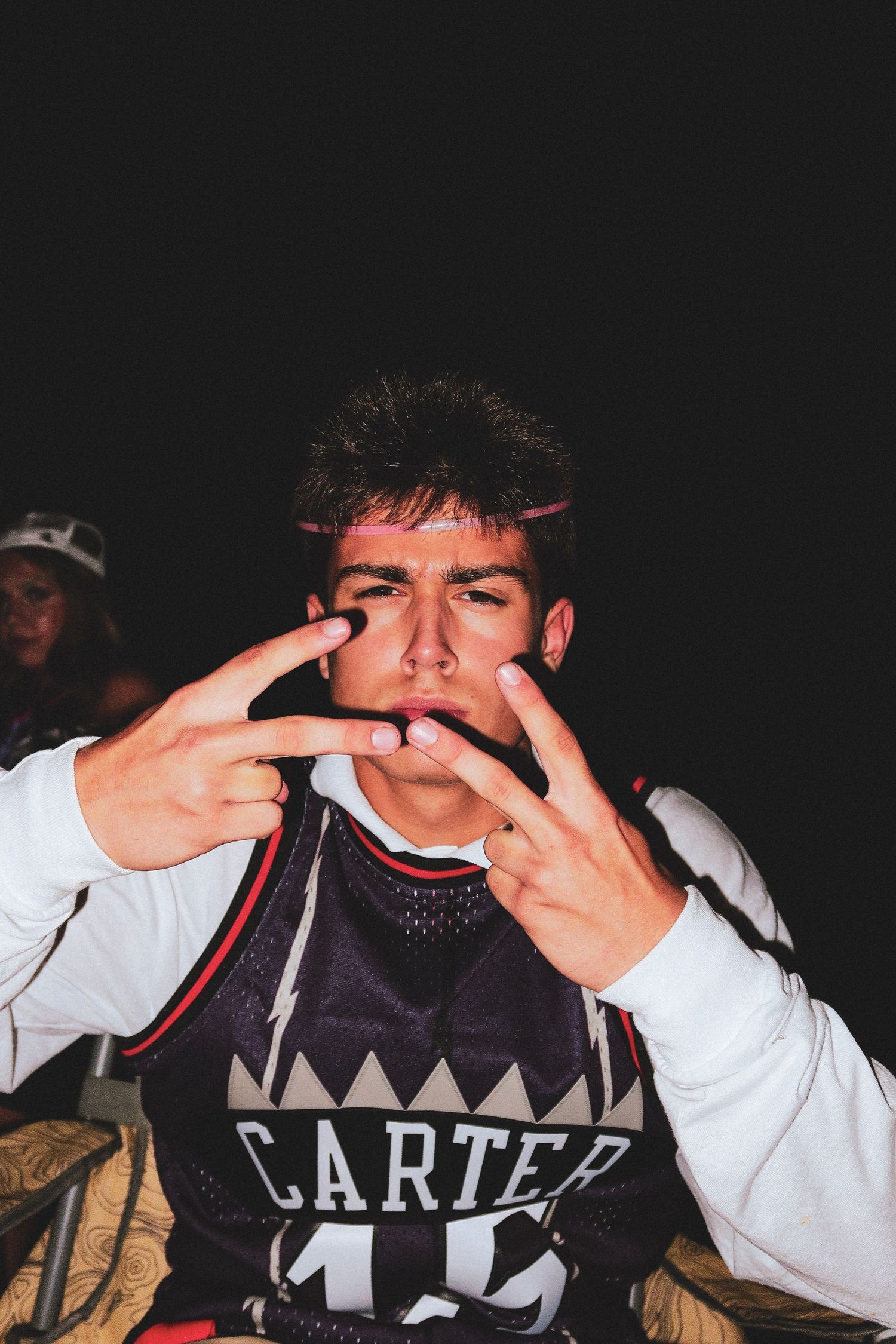 Young man in Carter jersey making hand gestures toward camera in a dark setting.
