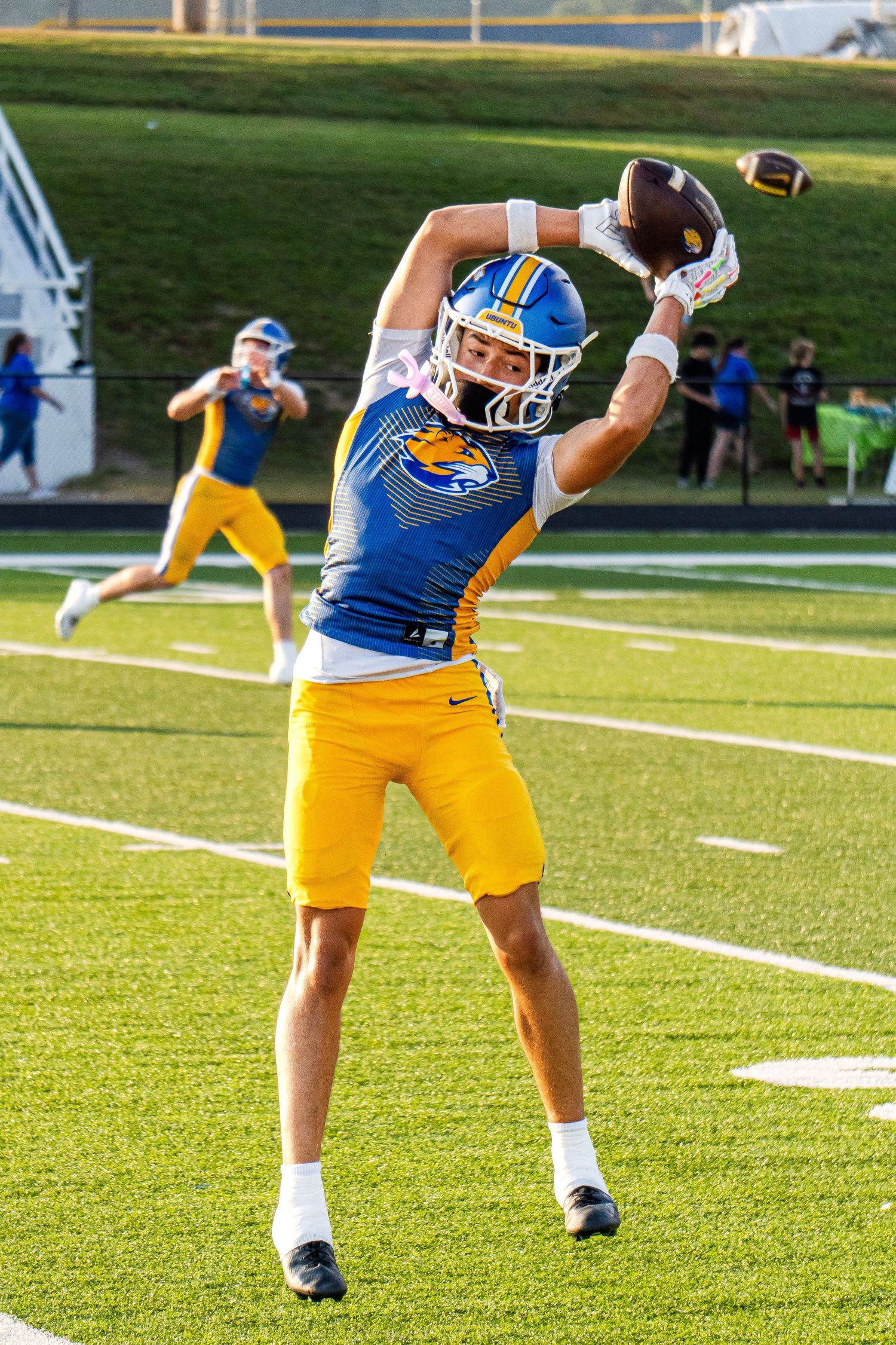 High school football player in blue and gold uniform catches a pass during warm-ups on the field.