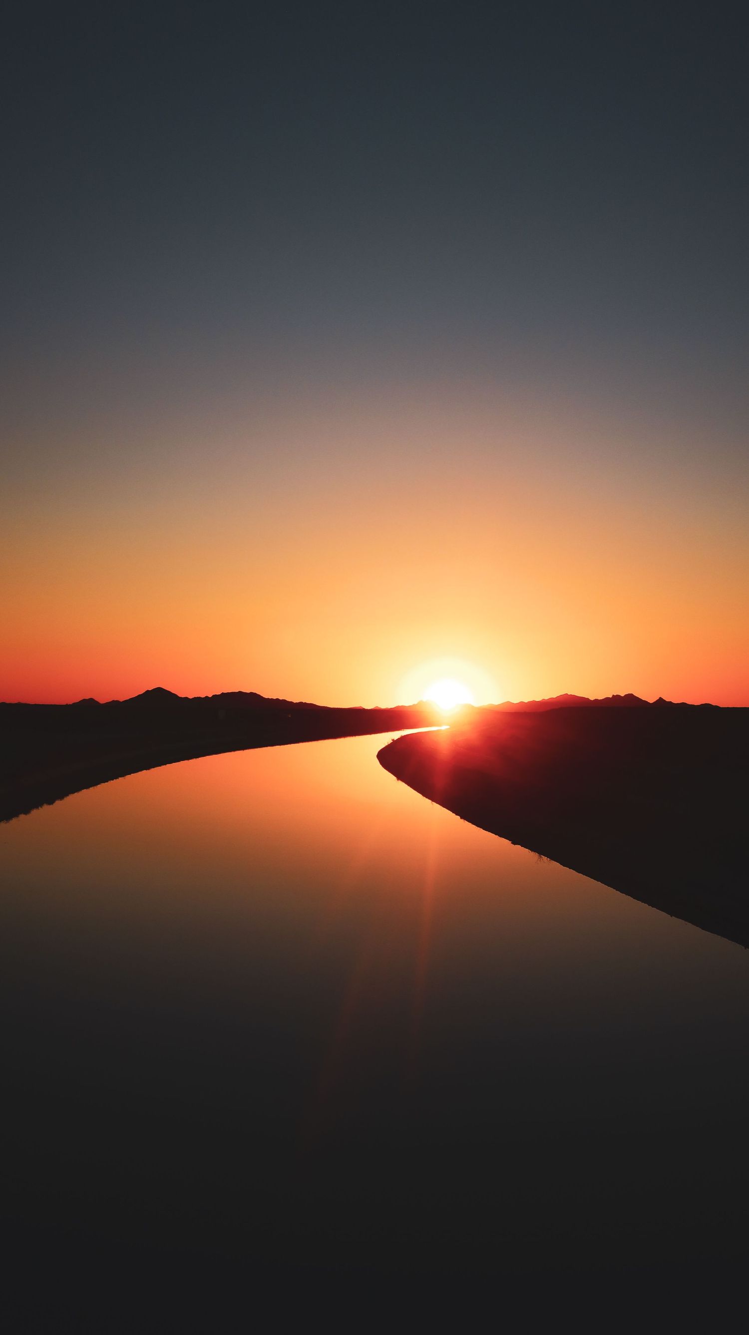 Stunning sunset over a calm river with silhouetted mountains and vibrant orange sky reflecting on still water.