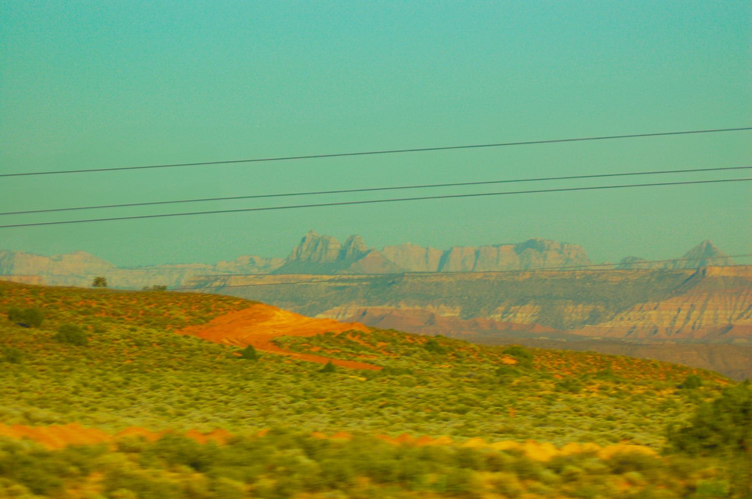 Desert landscape with red rock formations and green scrubland viewed through a car window under hazy sky.