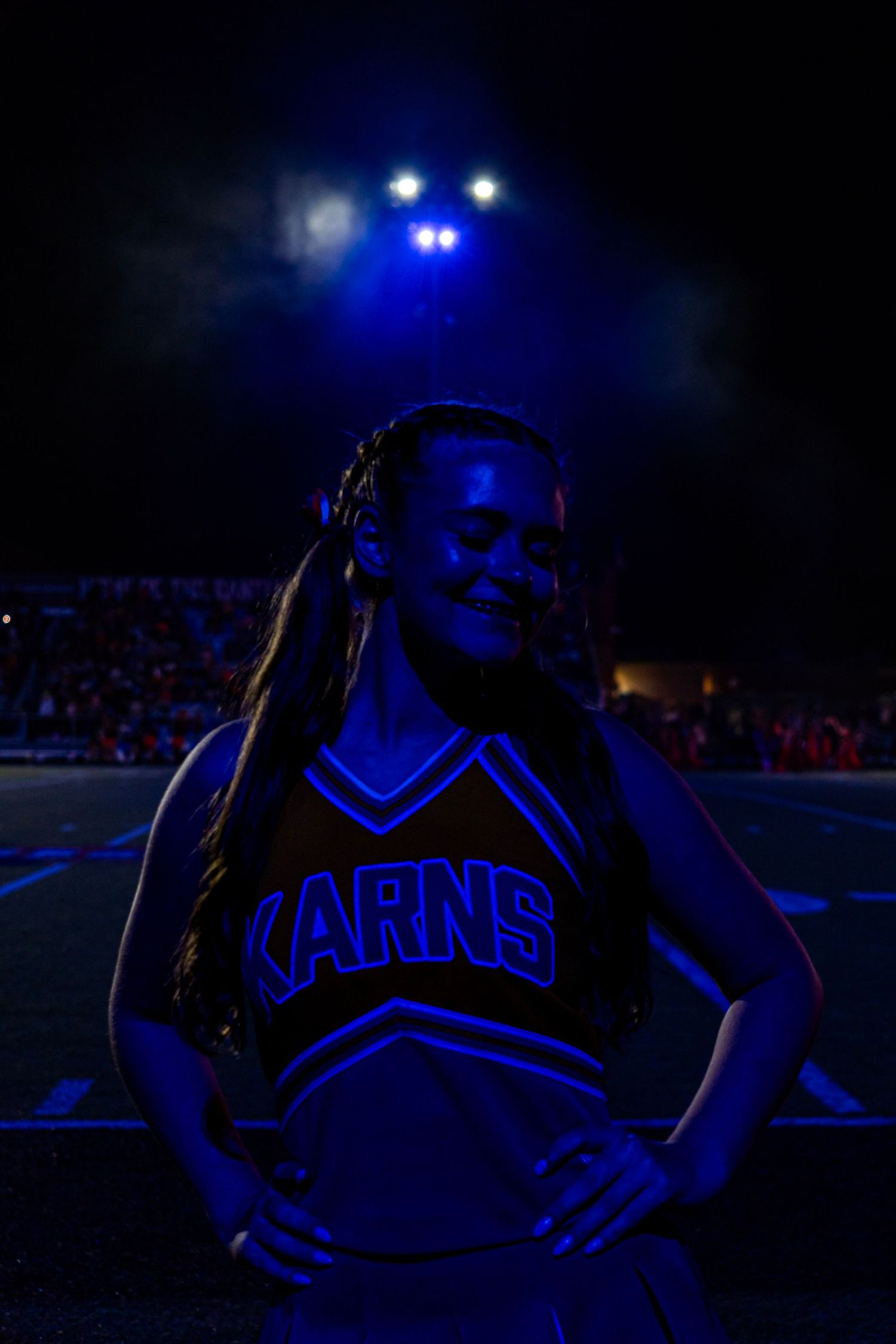 Karns cheerleader smiling under dramatic blue stadium lights at a night football game.