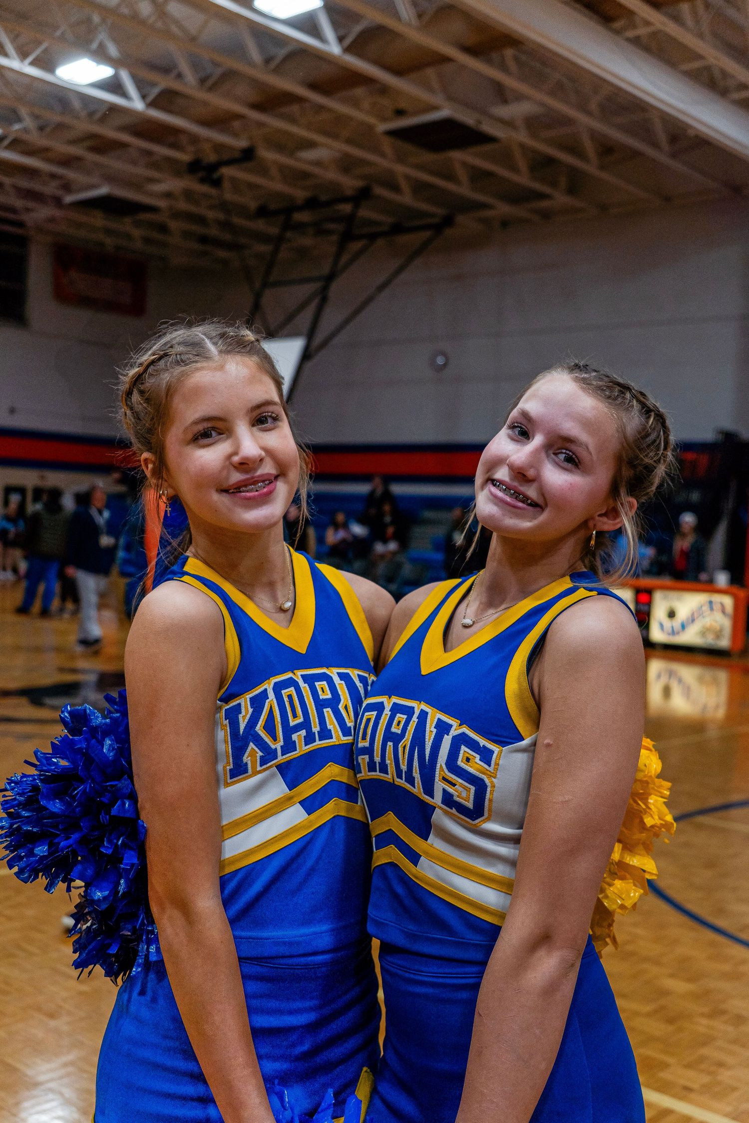 Two Karns cheerleaders smiling and posing together with blue pom-poms at a school basketball game.