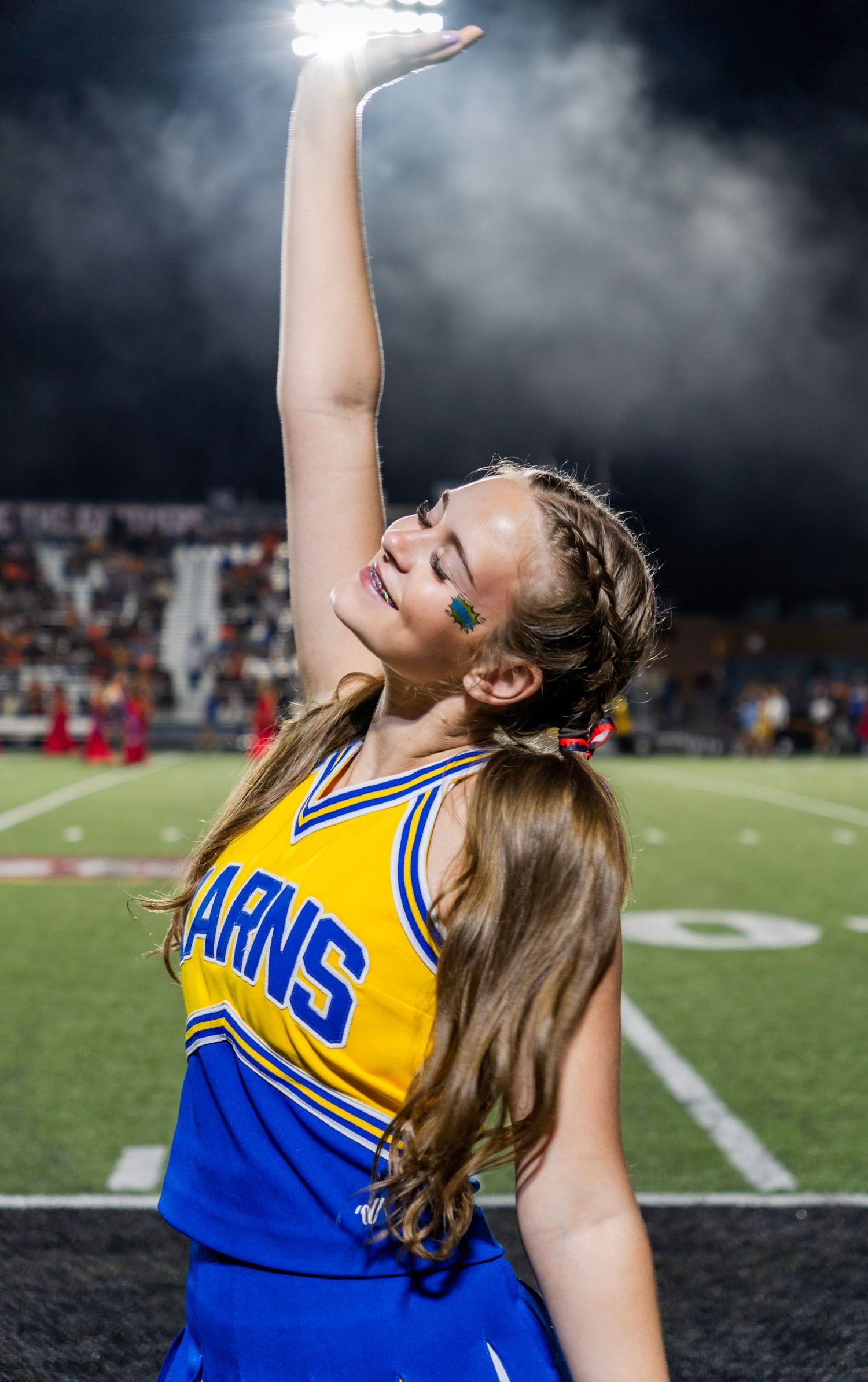 Karns cheerleader in yellow and blue uniform reaching arm up under bright stadium lights.
