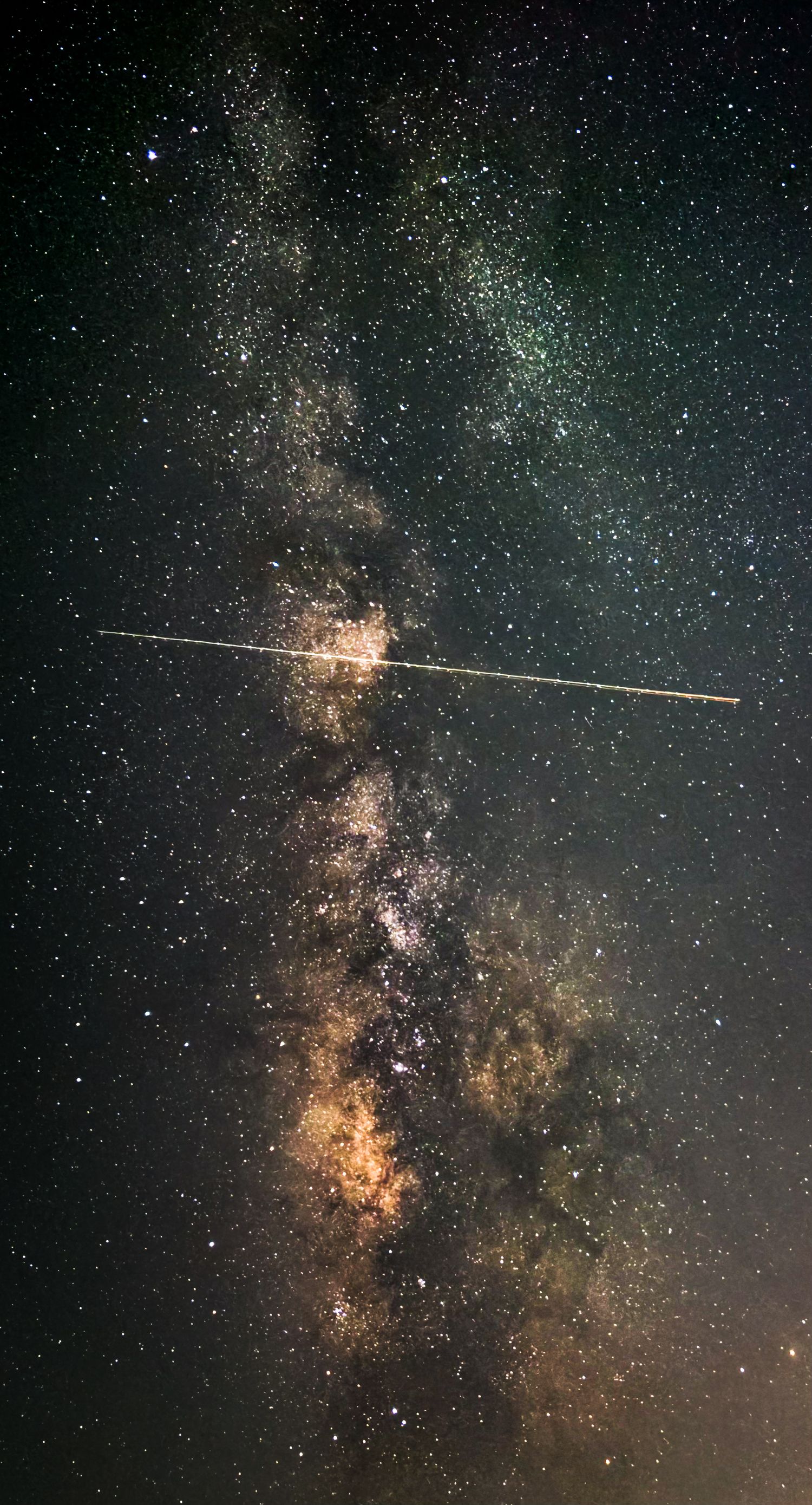 Milky Way galaxy with golden core and a bright satellite trail crossing the starry night sky.