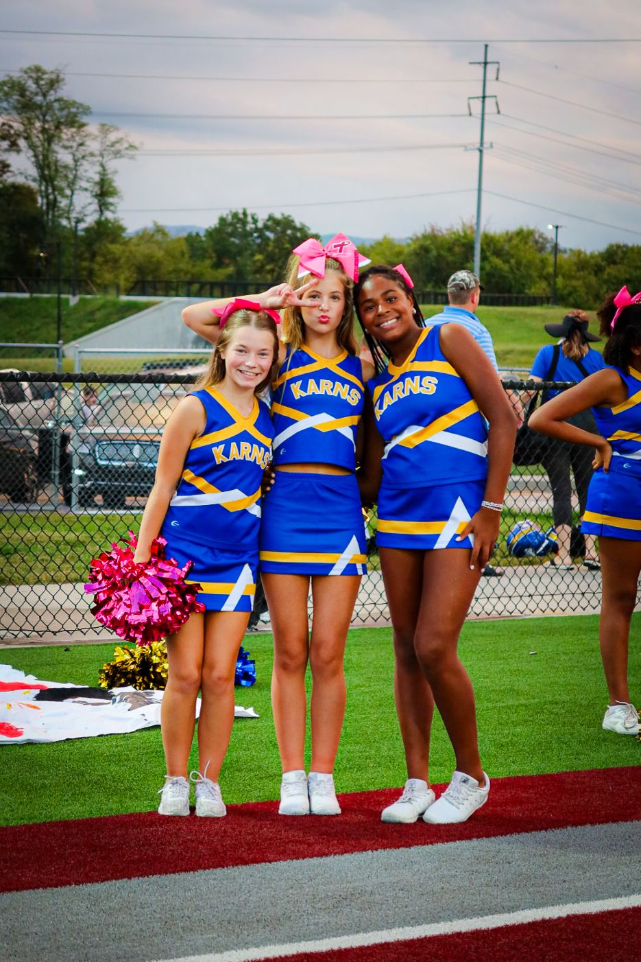 Three Karns cheerleaders in blue and gold uniforms posing together on a football field sideline.