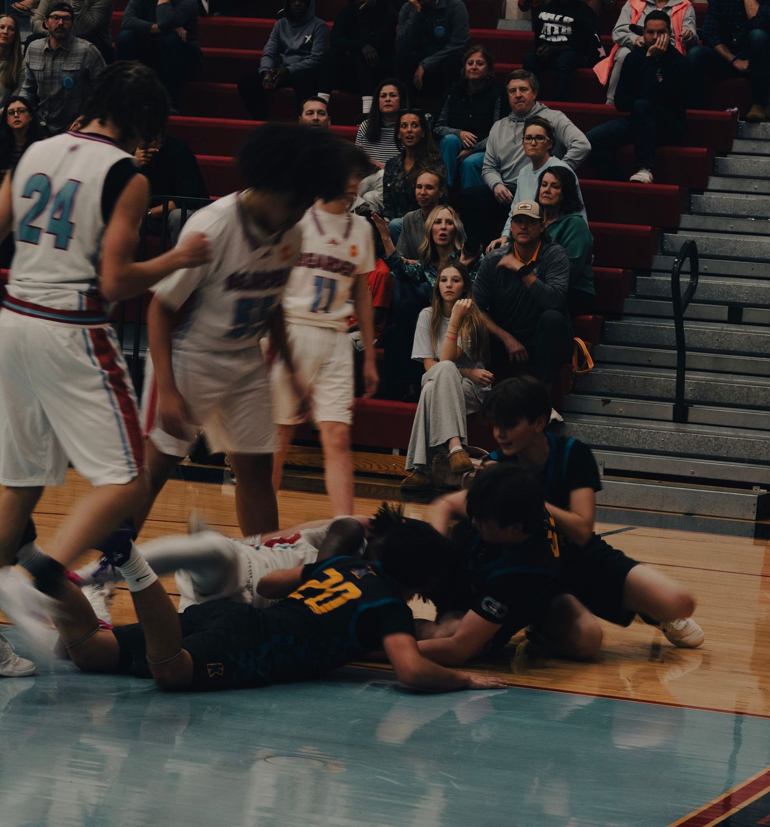 Players dive for a loose ball during an intense high school basketball game with fans watching.