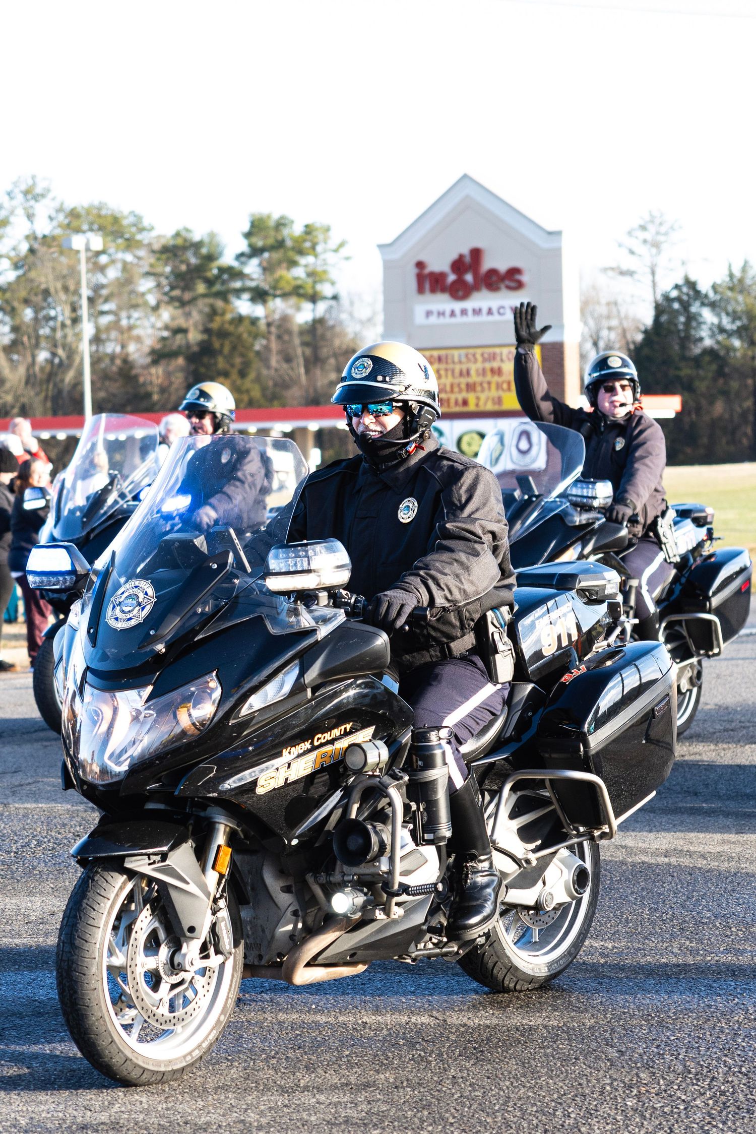 Police officers on black motorcycles waving while escorting a holiday parade past Ingles pharmacy.