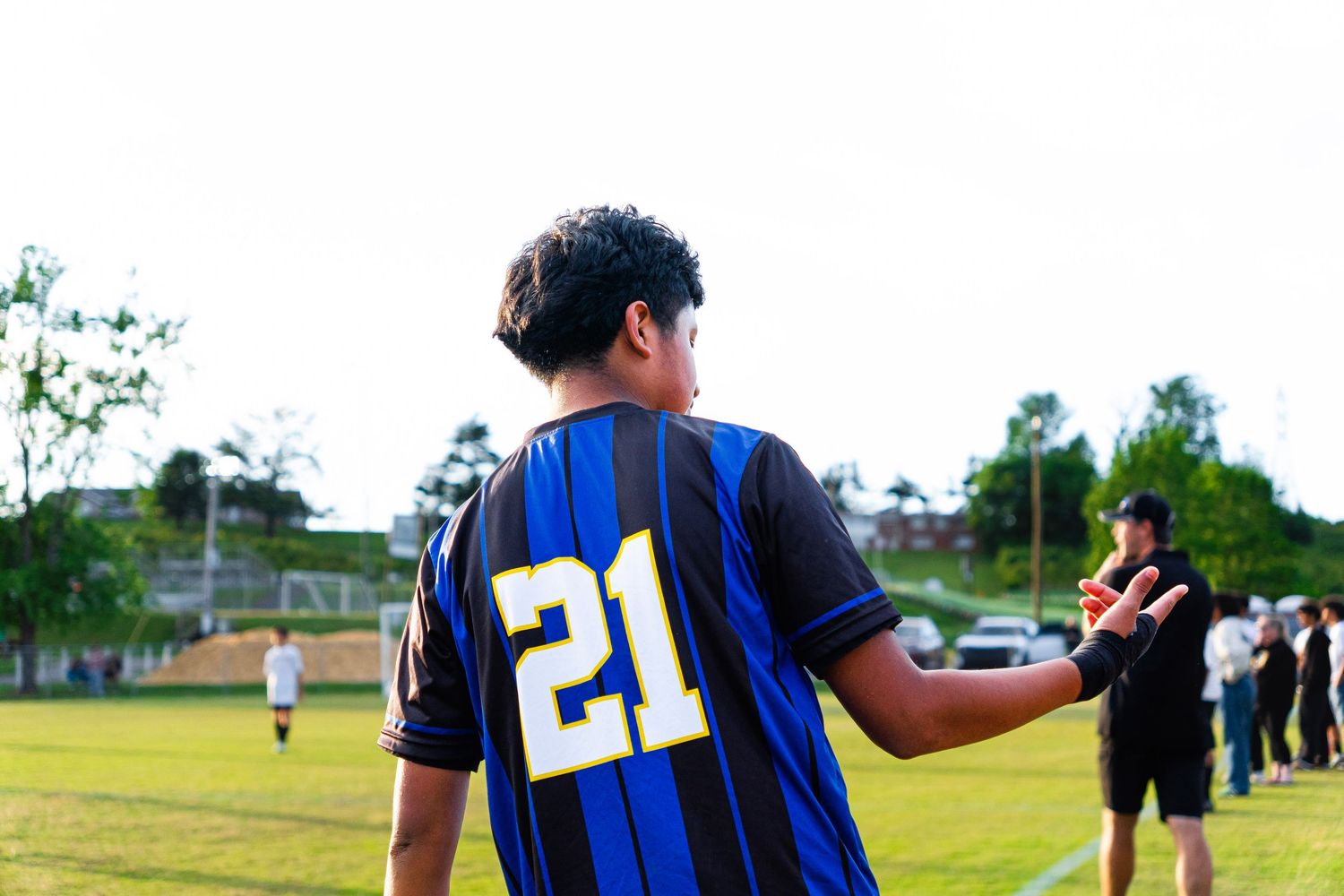Youth soccer player wearing #21 blue striped jersey gestures on green field during game.