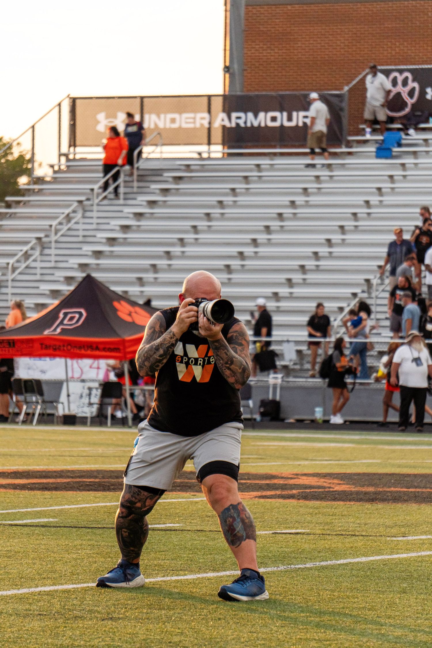 A tattooed photographer crouches on a football field capturing game action with a telephoto lens.