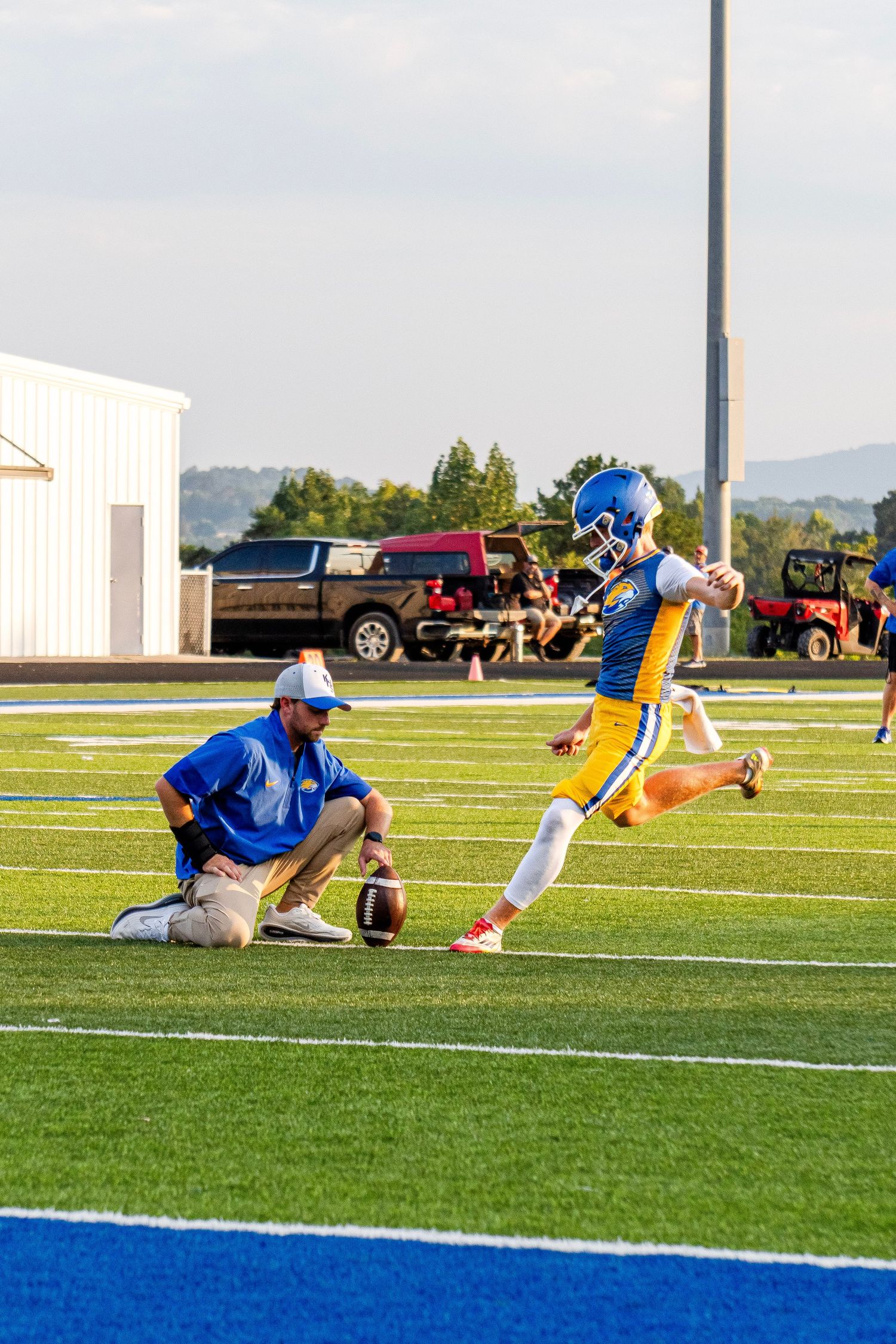 Football player practices a field goal kick while coach holds the ball on a blue turf field.