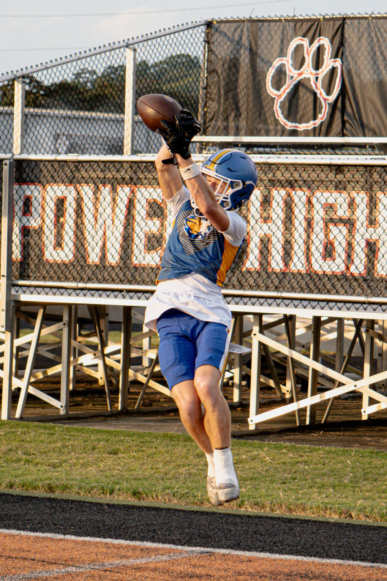 A football player in blue uniform leaps to catch a pass during practice at Powell High School stadium.