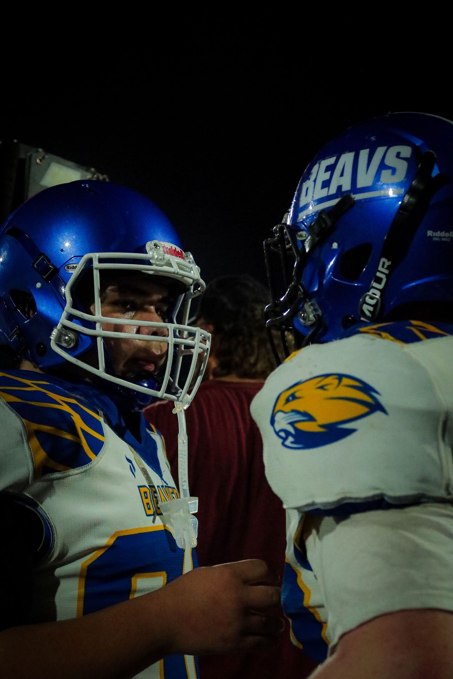 Two football players in blue Beavs helmets facing each other intensely during a night game.