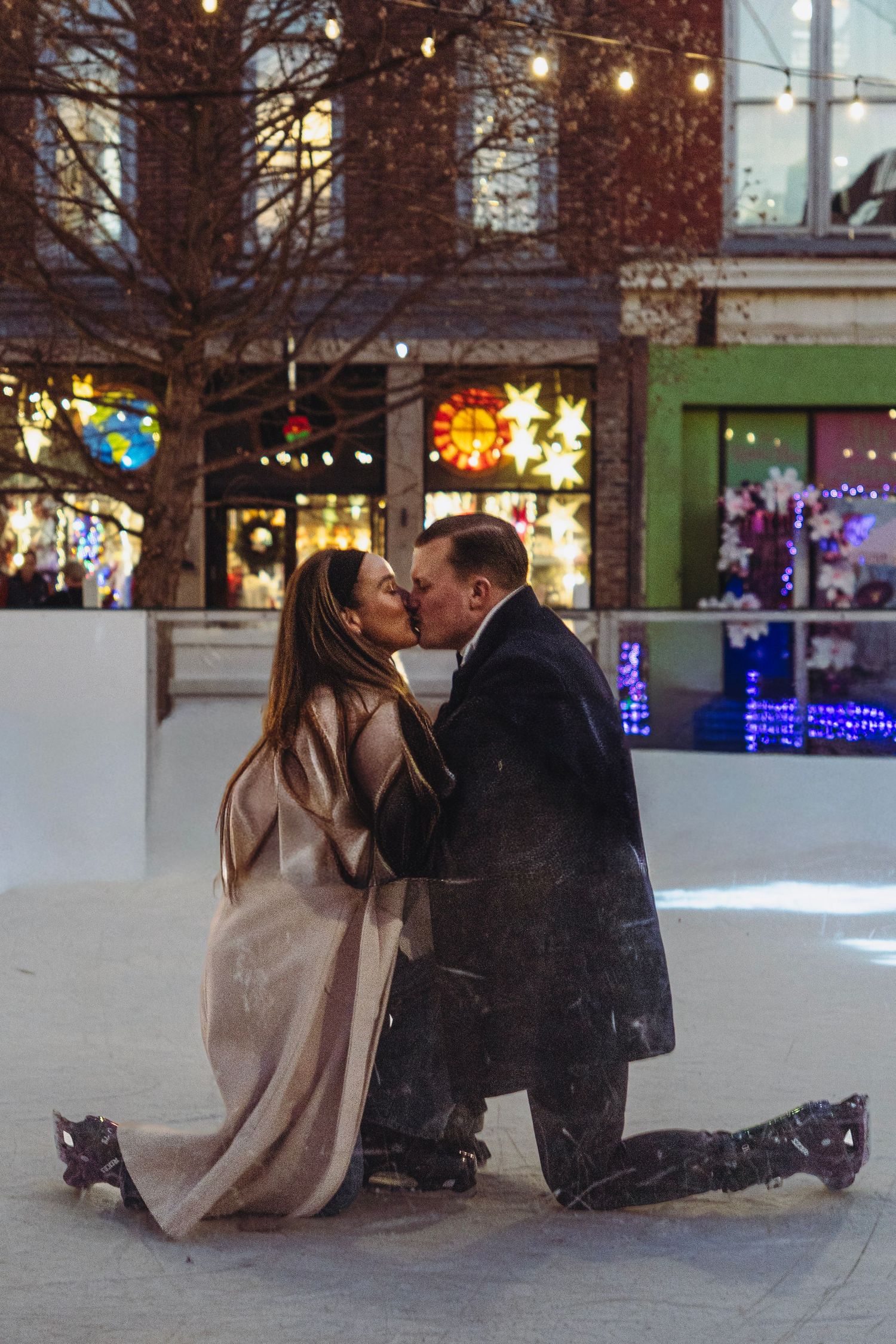 Couple sharing a kiss on their knees at an outdoor ice skating rink with holiday lights.