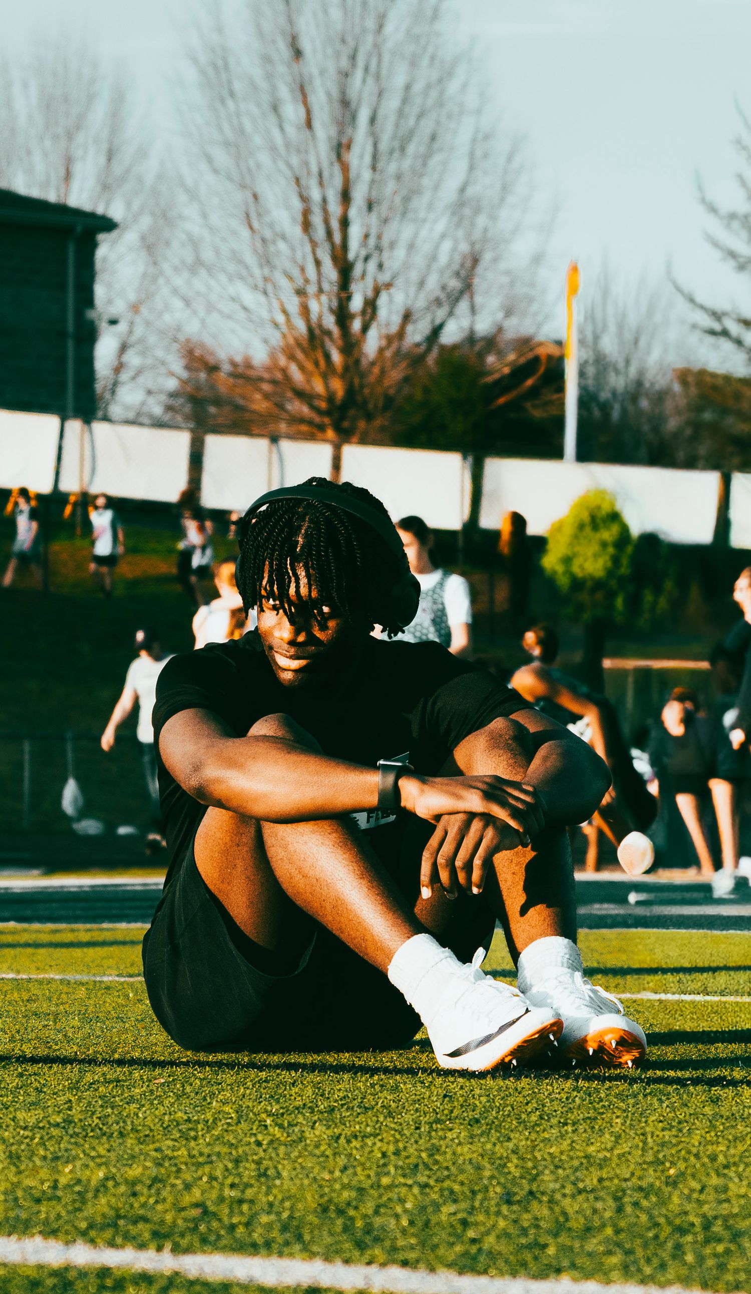 Male athlete with dreadlocks sits on green turf field hugging knees during outdoor sports event.