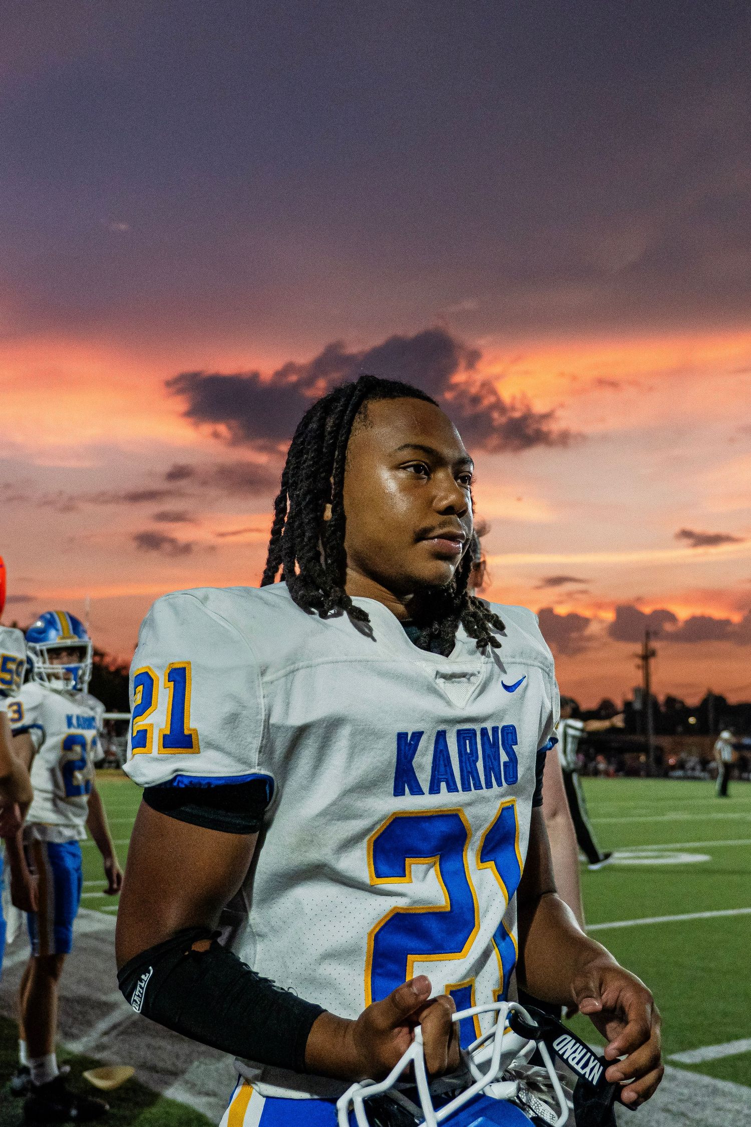 Karns football player number 21 stands on the sideline with a dramatic sunset sky behind him.