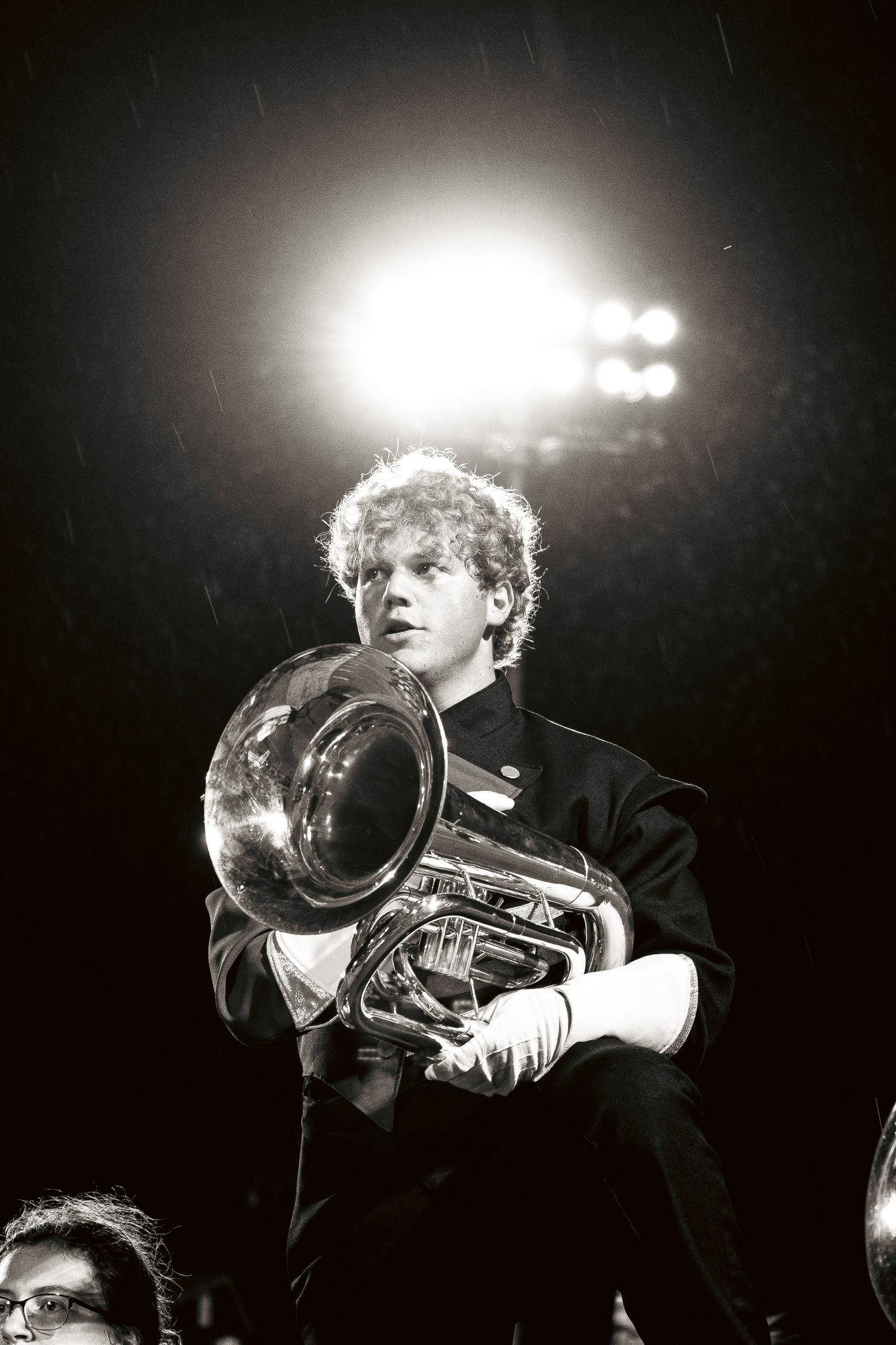 A marching band member plays a tuba in the rain under bright stadium lights at night.