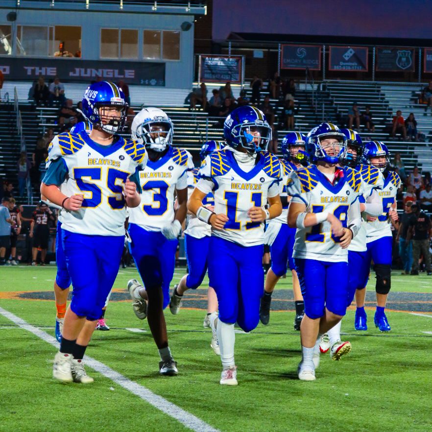 High school football team in blue uniforms runs onto field under stadium lights.