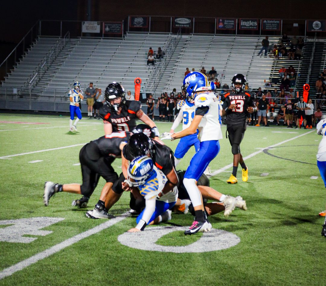 High school football players in black and blue uniforms collide during an intense tackle on a night game field.