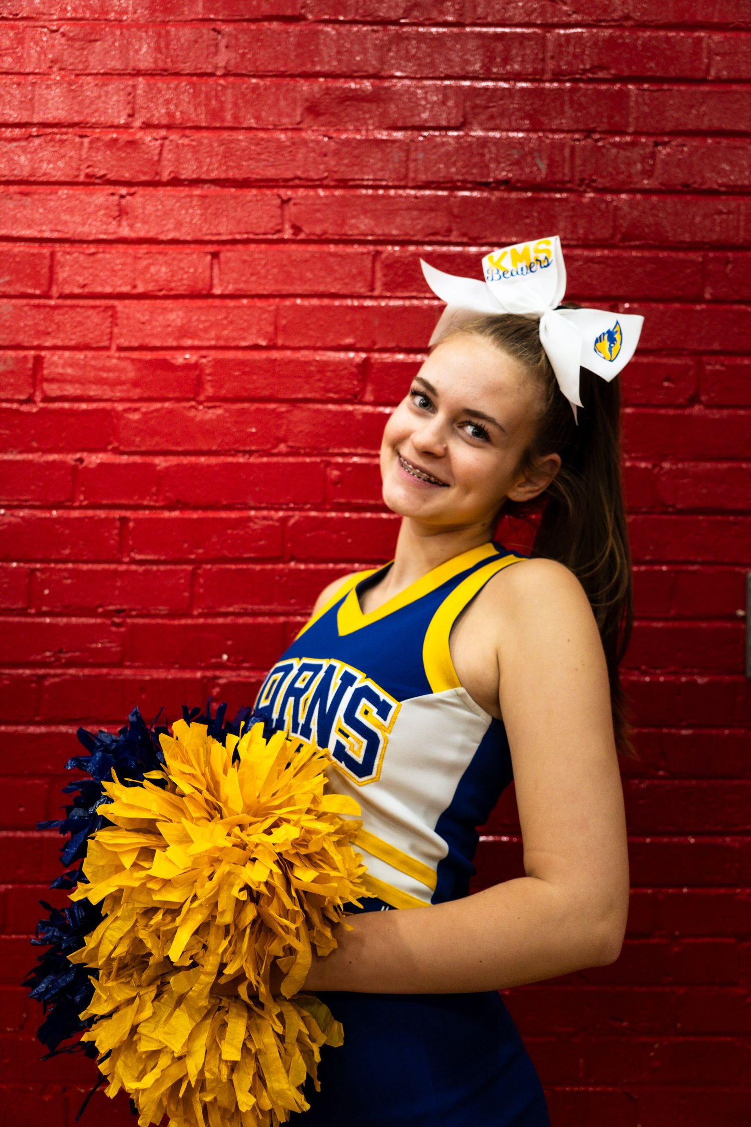 Karns cheerleader in blue and gold uniform poses with pom-poms against red brick wall.