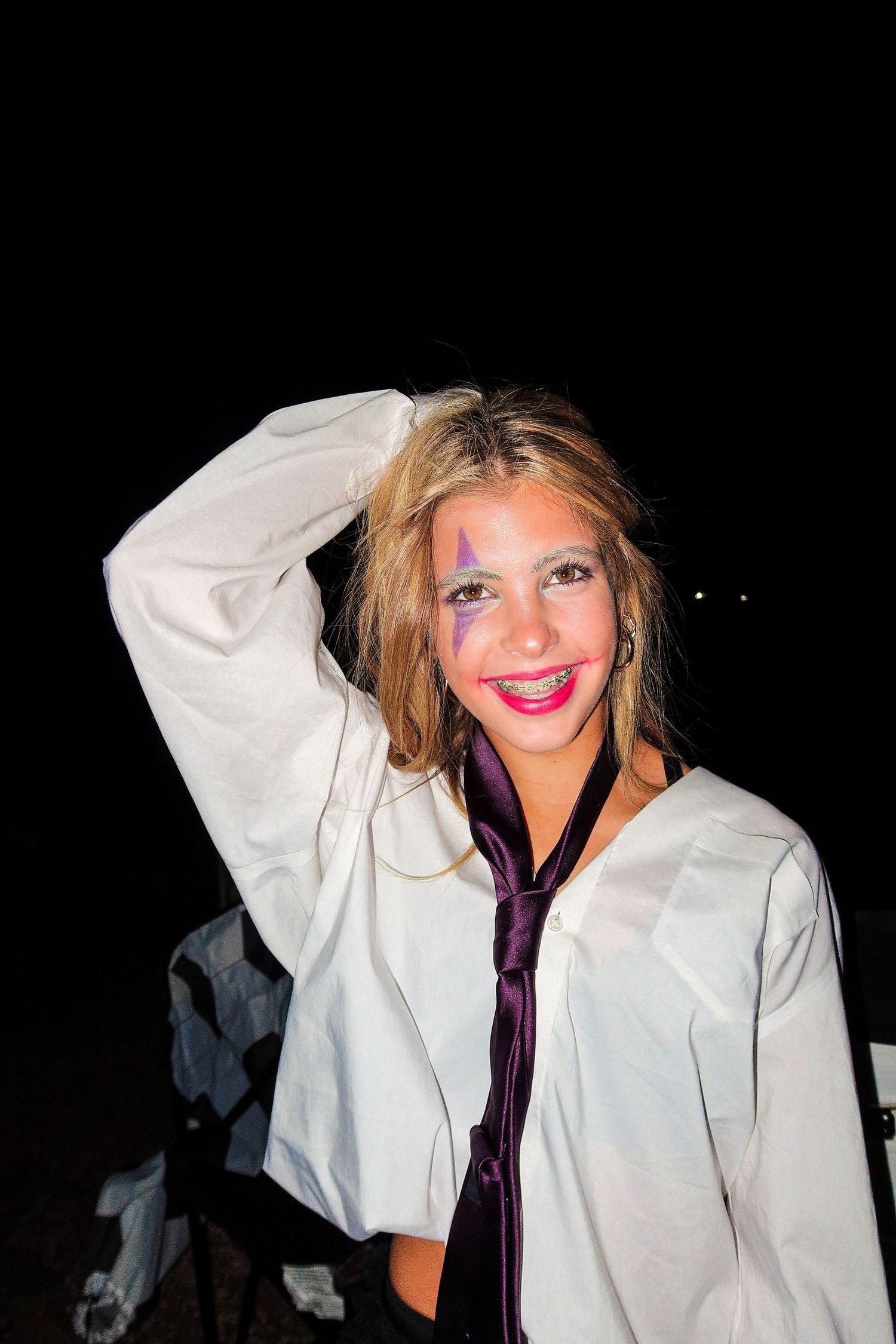 Smiling teenage girl in white shirt and tie with colorful face paint posing outdoors at night.