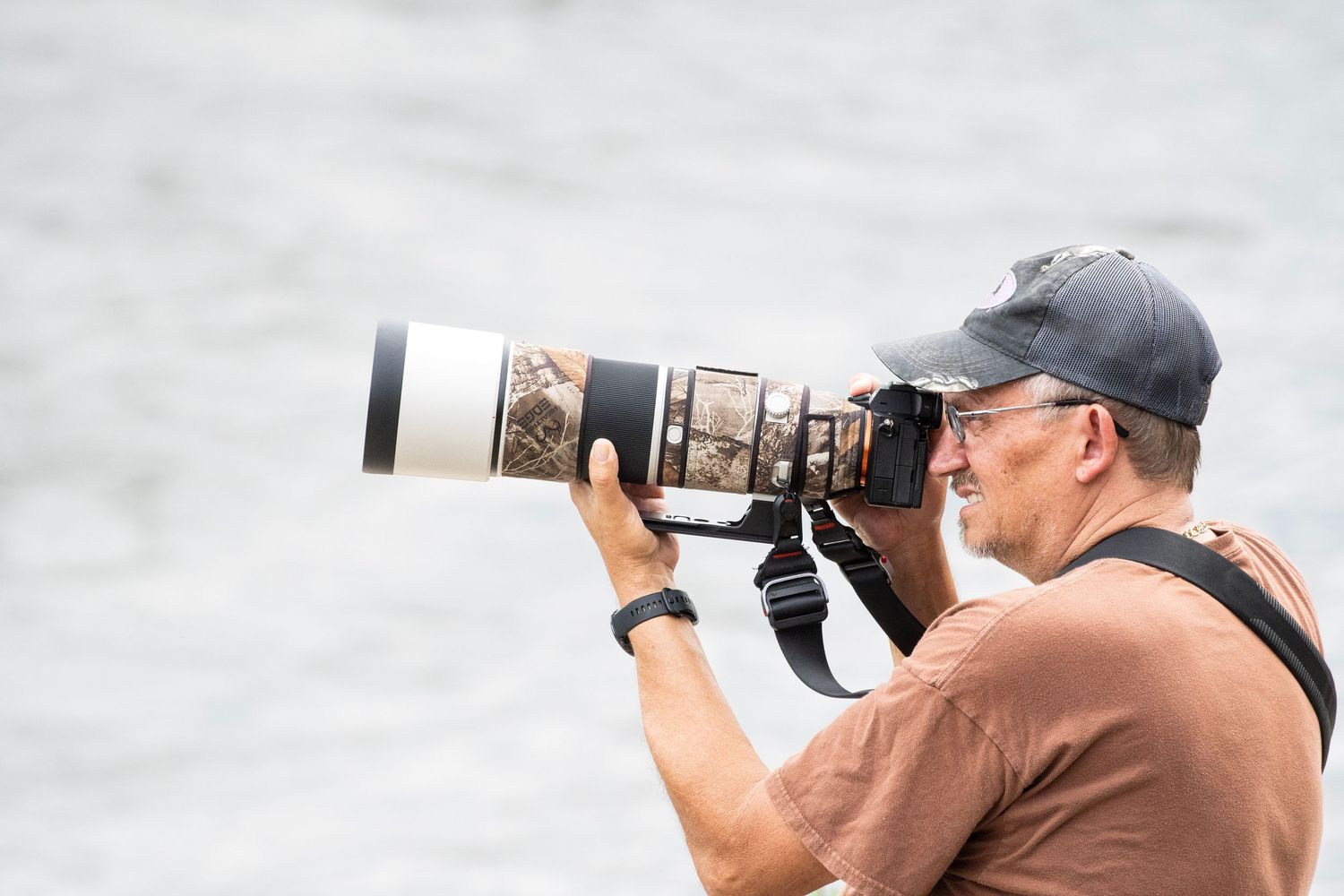 Male wildlife photographer using large telephoto lens camera outdoors near water.