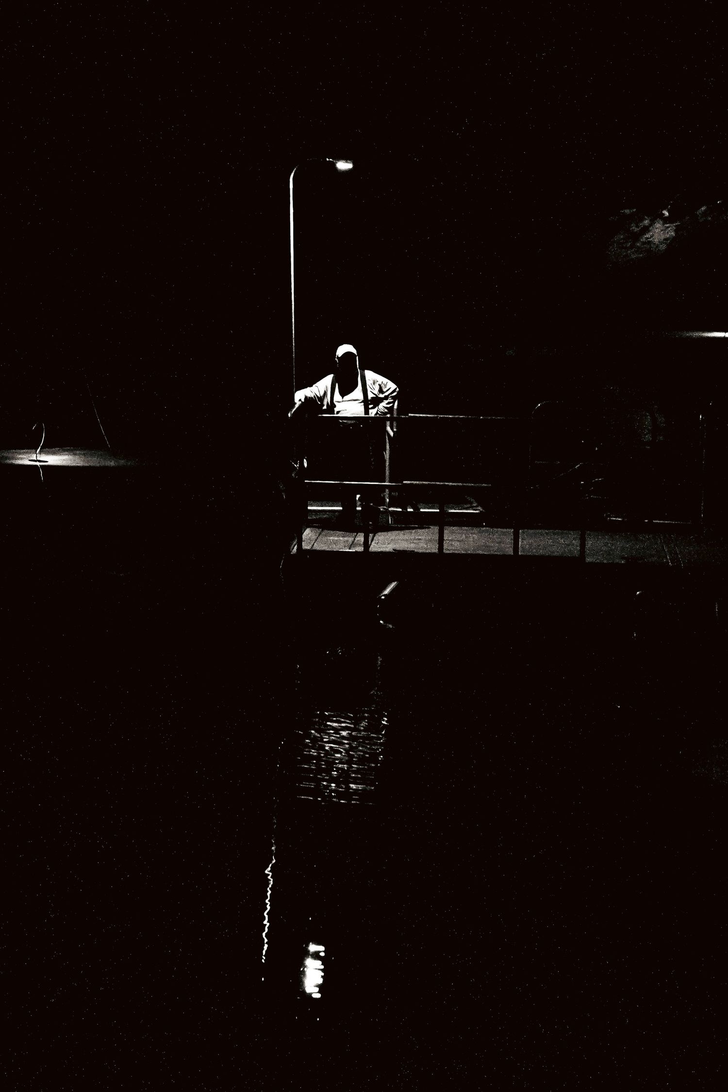 Silhouette of a person standing on a bridge at night under a street lamp with water reflection.