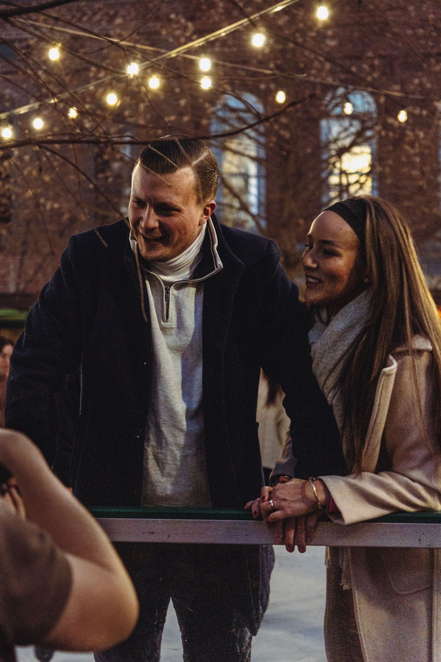Smiling couple leaning on a railing together outdoors at night under glowing string lights.
