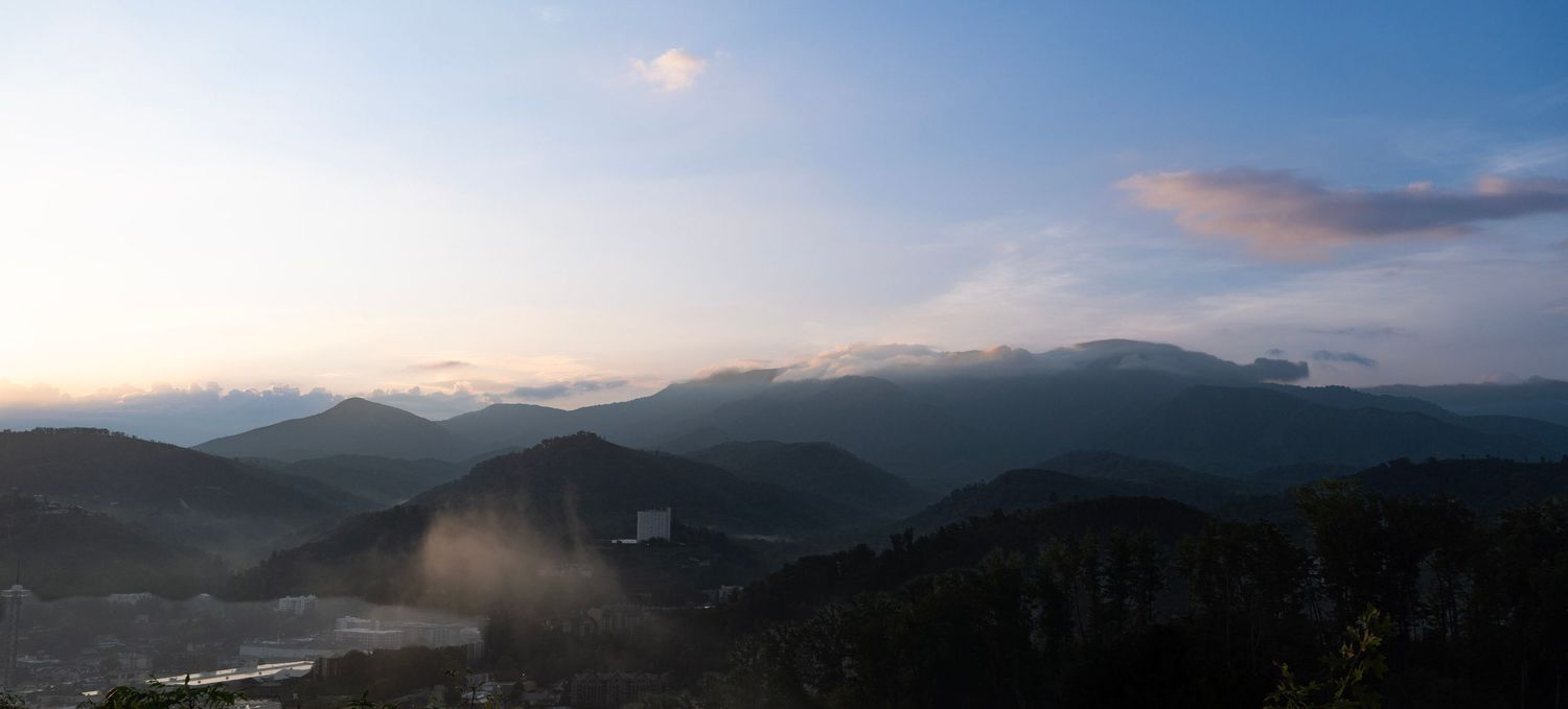 Panoramic view of misty mountain ranges at dusk with soft blue sky and scattered clouds.