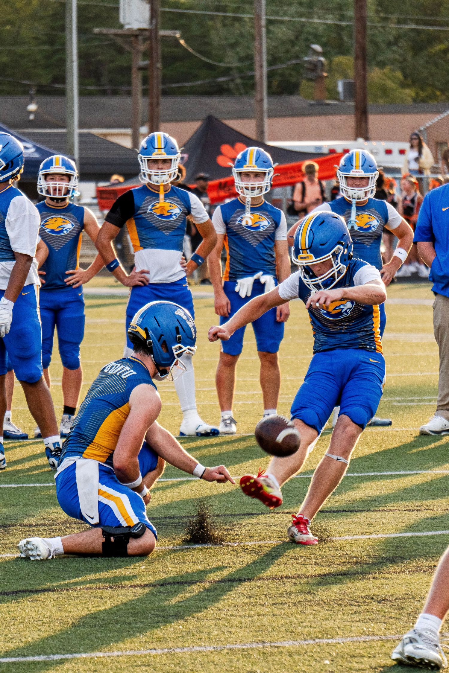 High school football players in blue and gold uniforms practice field goal kicks on the field.