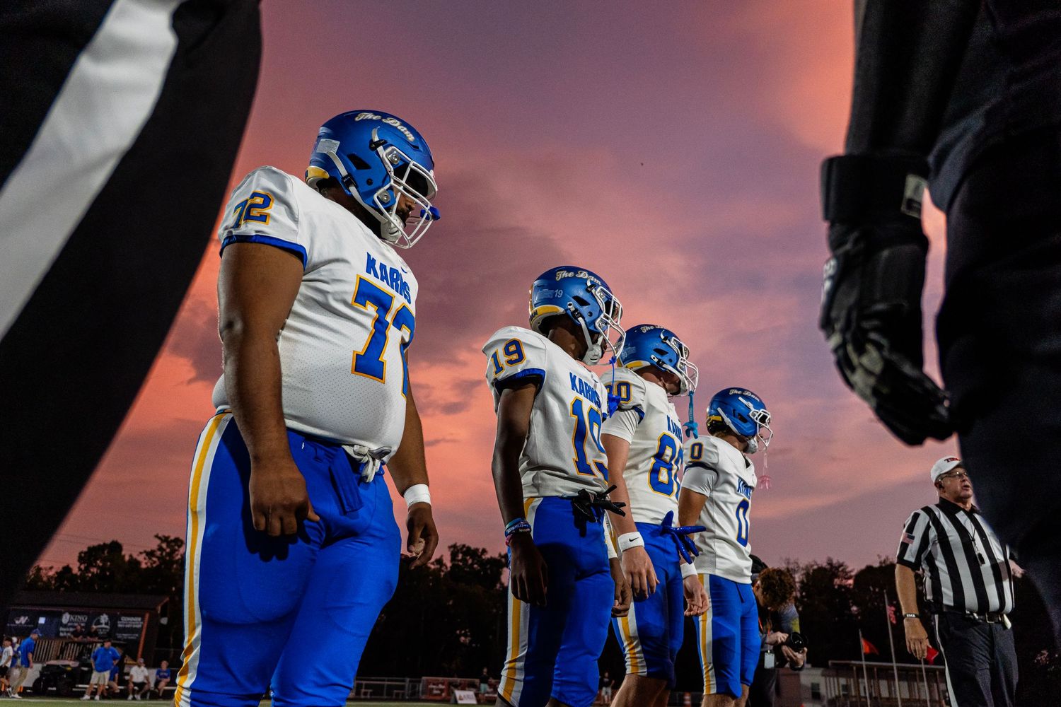 Karns football players in blue and gold uniforms lining up at sunset with a referee.