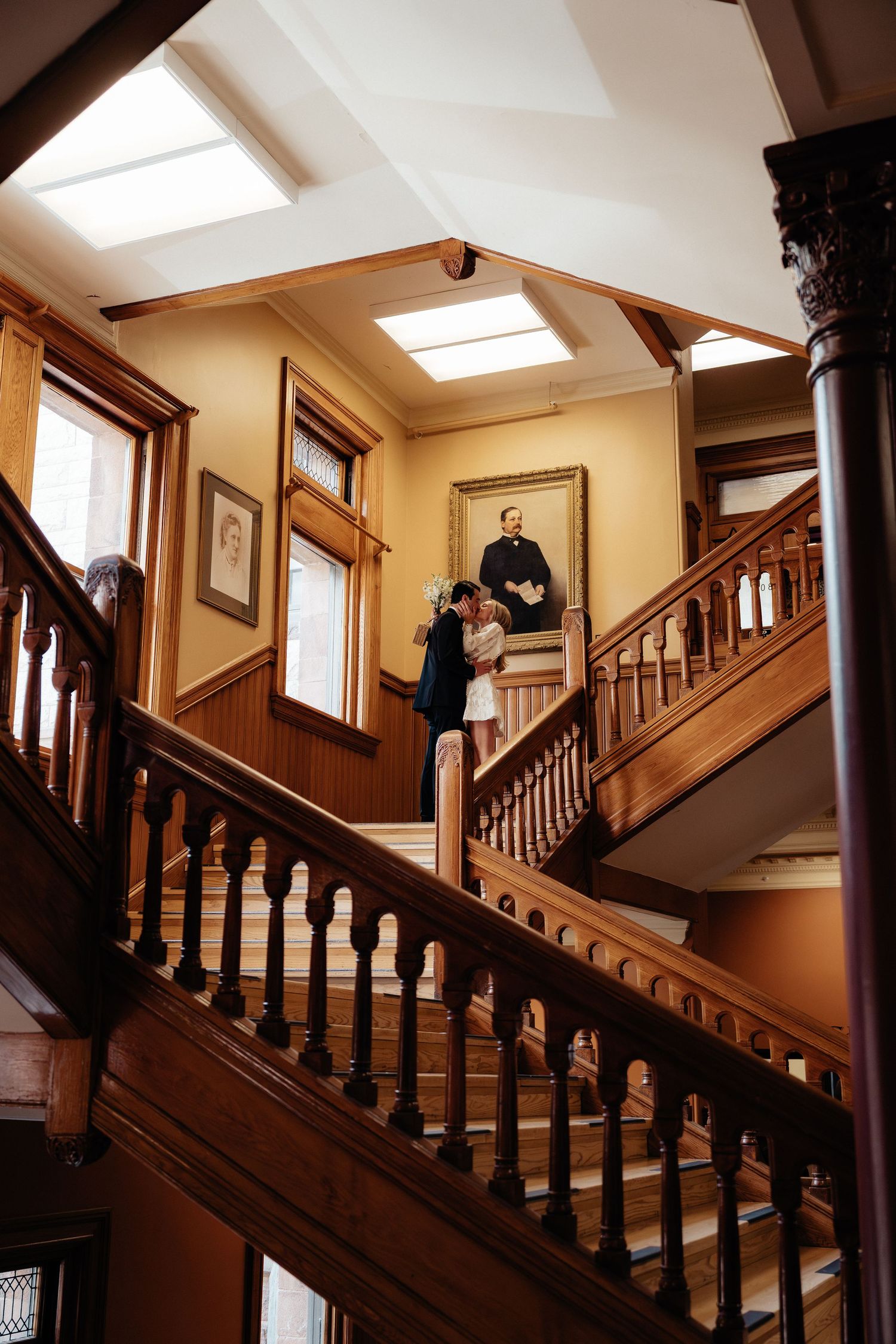A couple poses on a grand wooden staircase in a historic building with skylights.