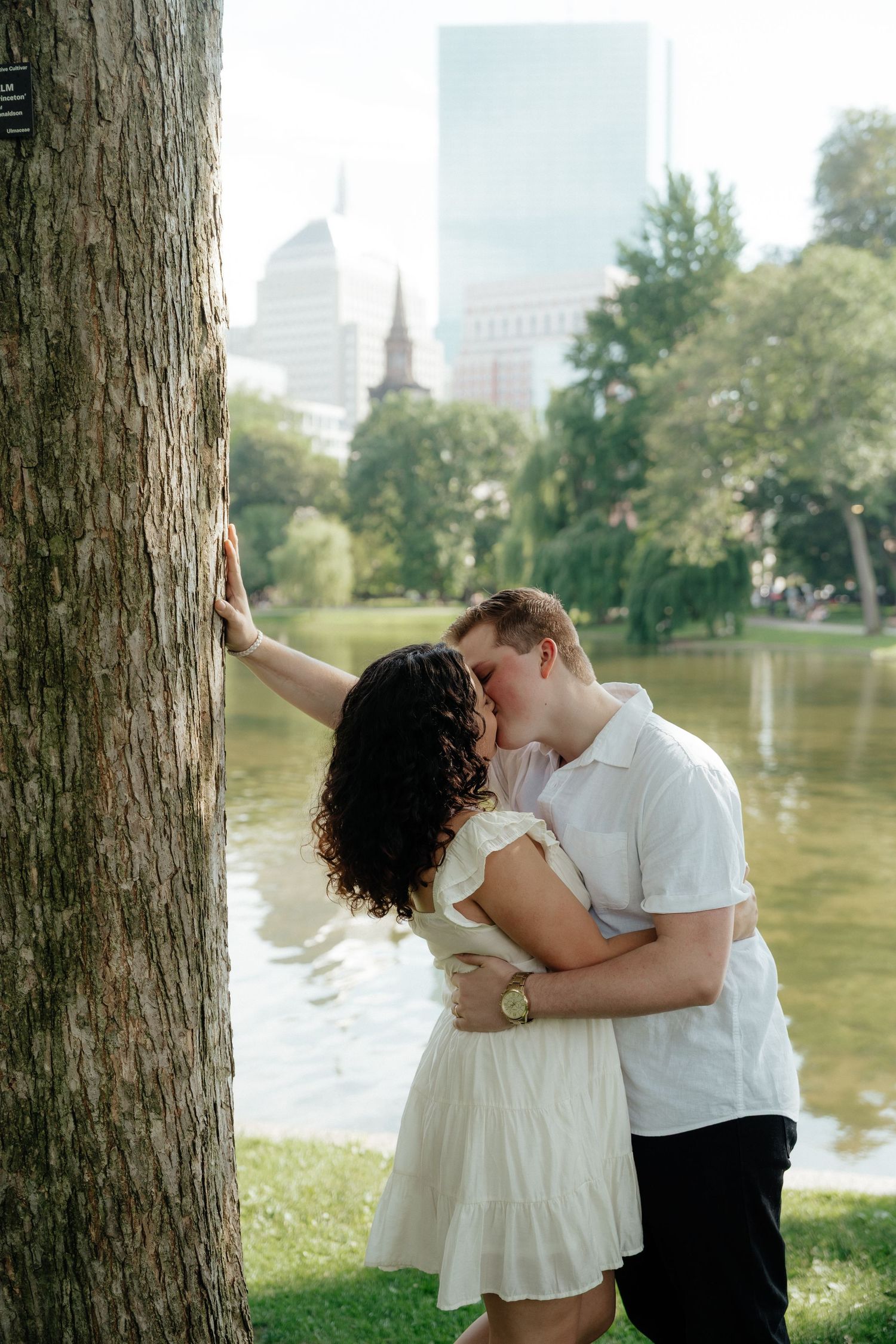 A tender moment shared against a large tree trunk with a pond and city skyline in the background.