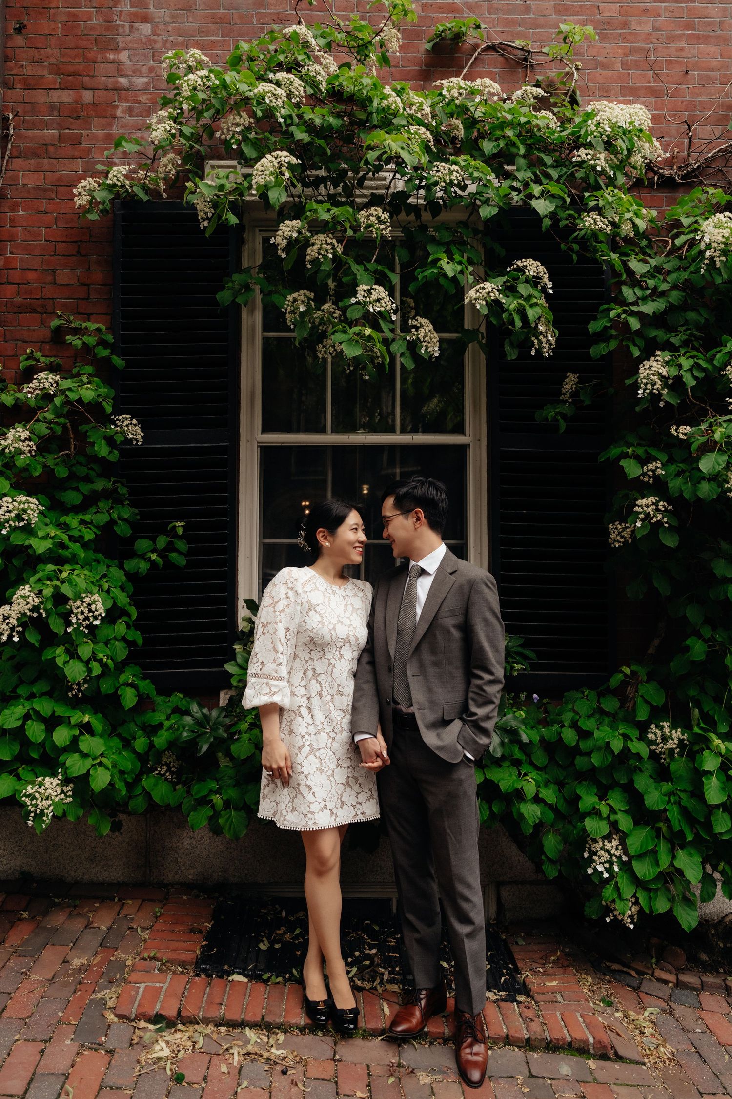 A couple stands against a brick wall with flowering vines creating a romantic garden setting.
