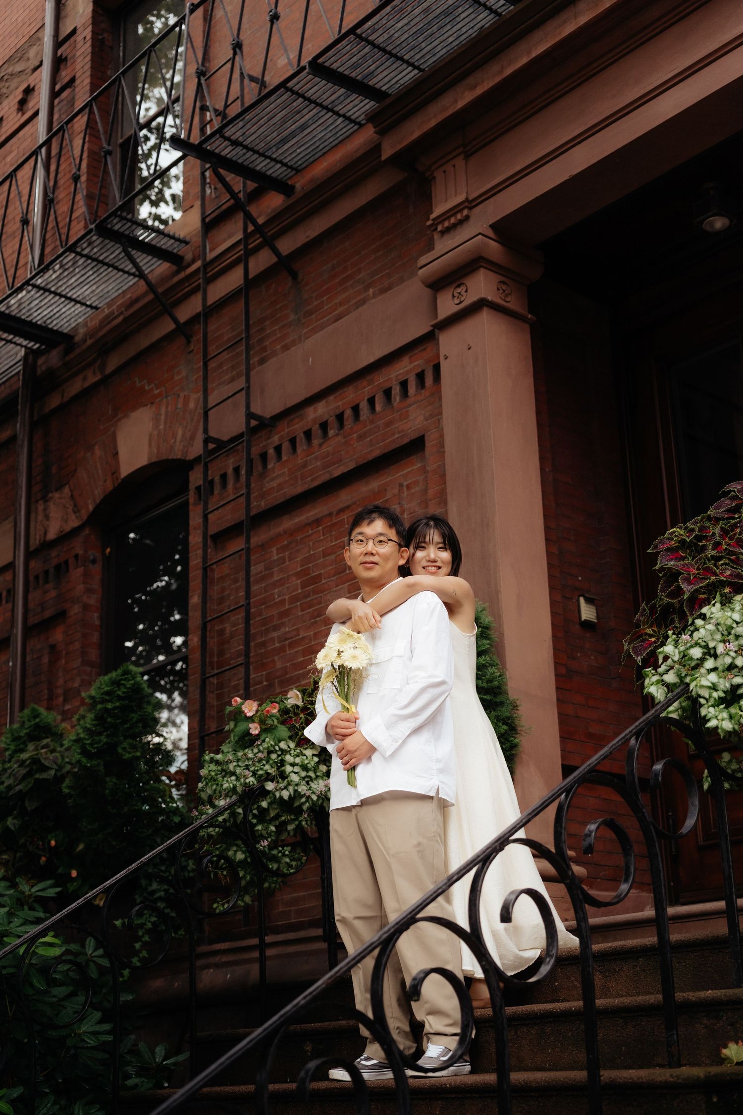 A couple embraces on decorative iron steps of a brownstone with hanging flower baskets.