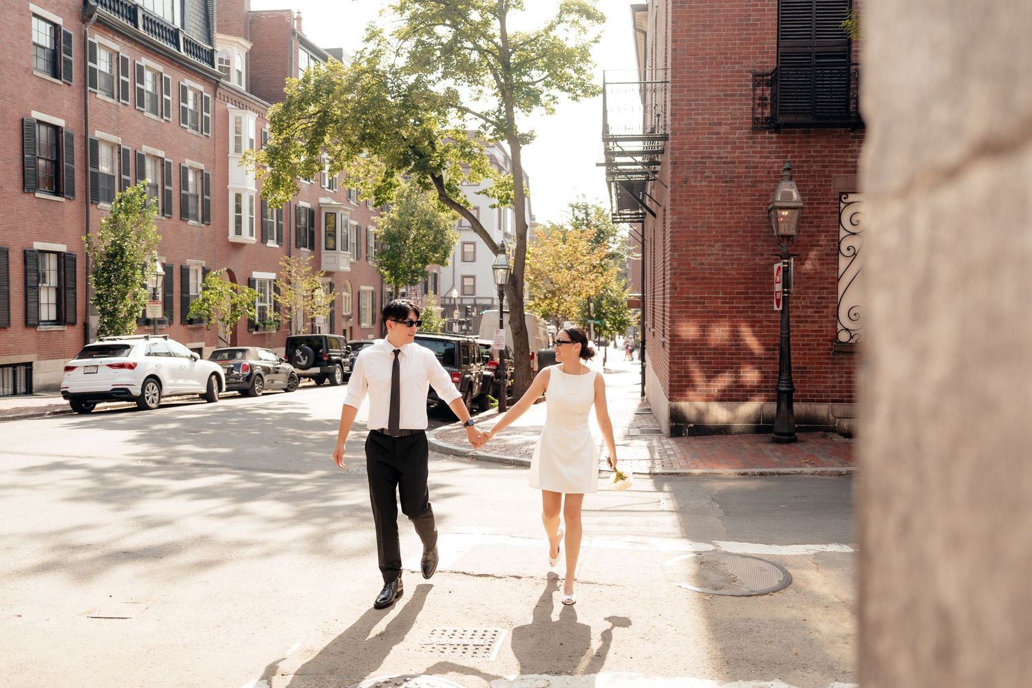 A couple walking hand in hand down a sunny Boston street lined with brick buildings.