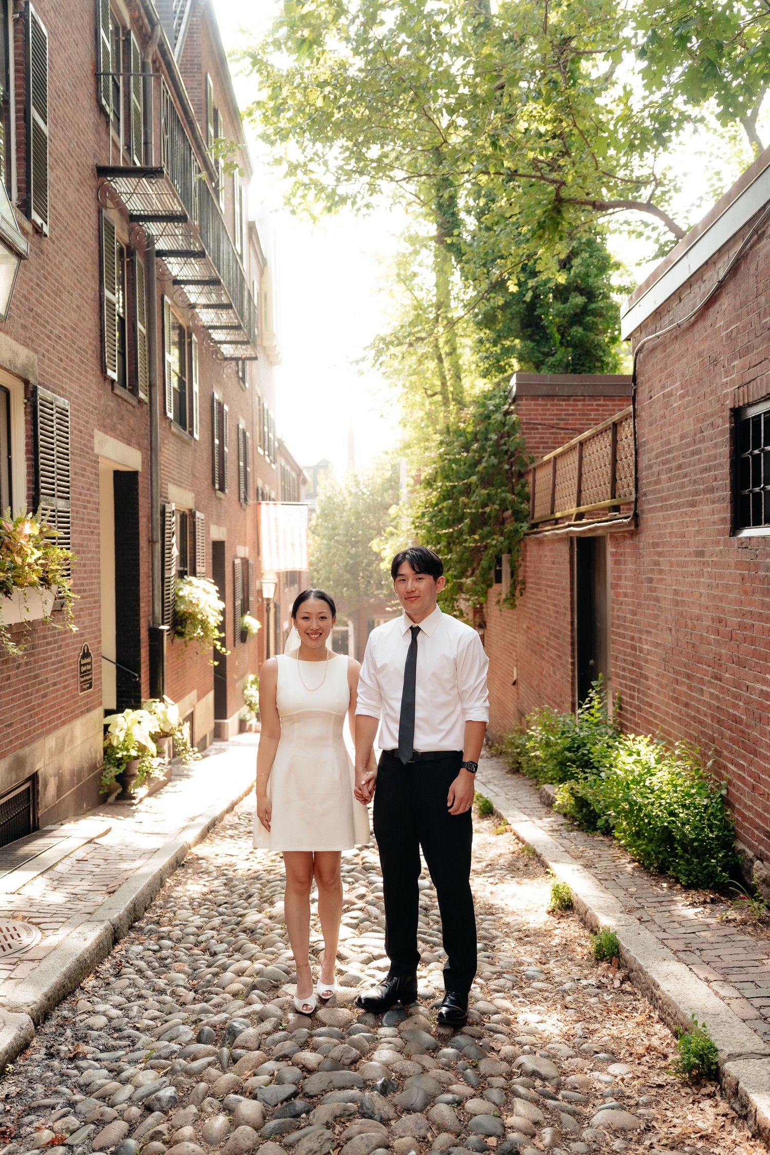 A couple in formal attire stands together on a historic cobblestone street in Boston's Beacon Hill neighborhood.