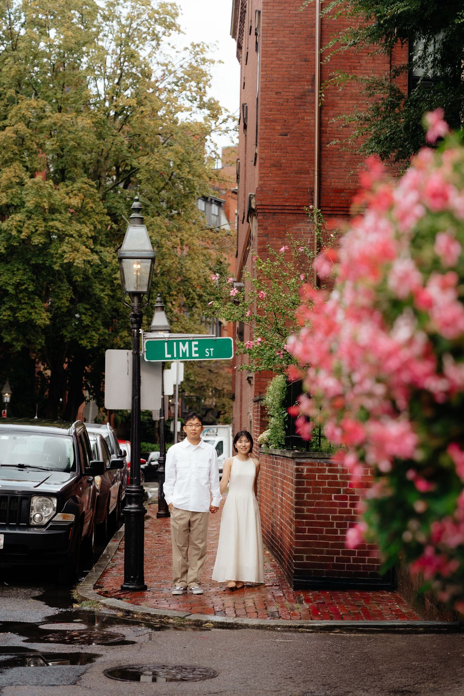 A couple stands on a brick sidewalk near a street sign for Lime St with pink flowers blooming in the foreground.