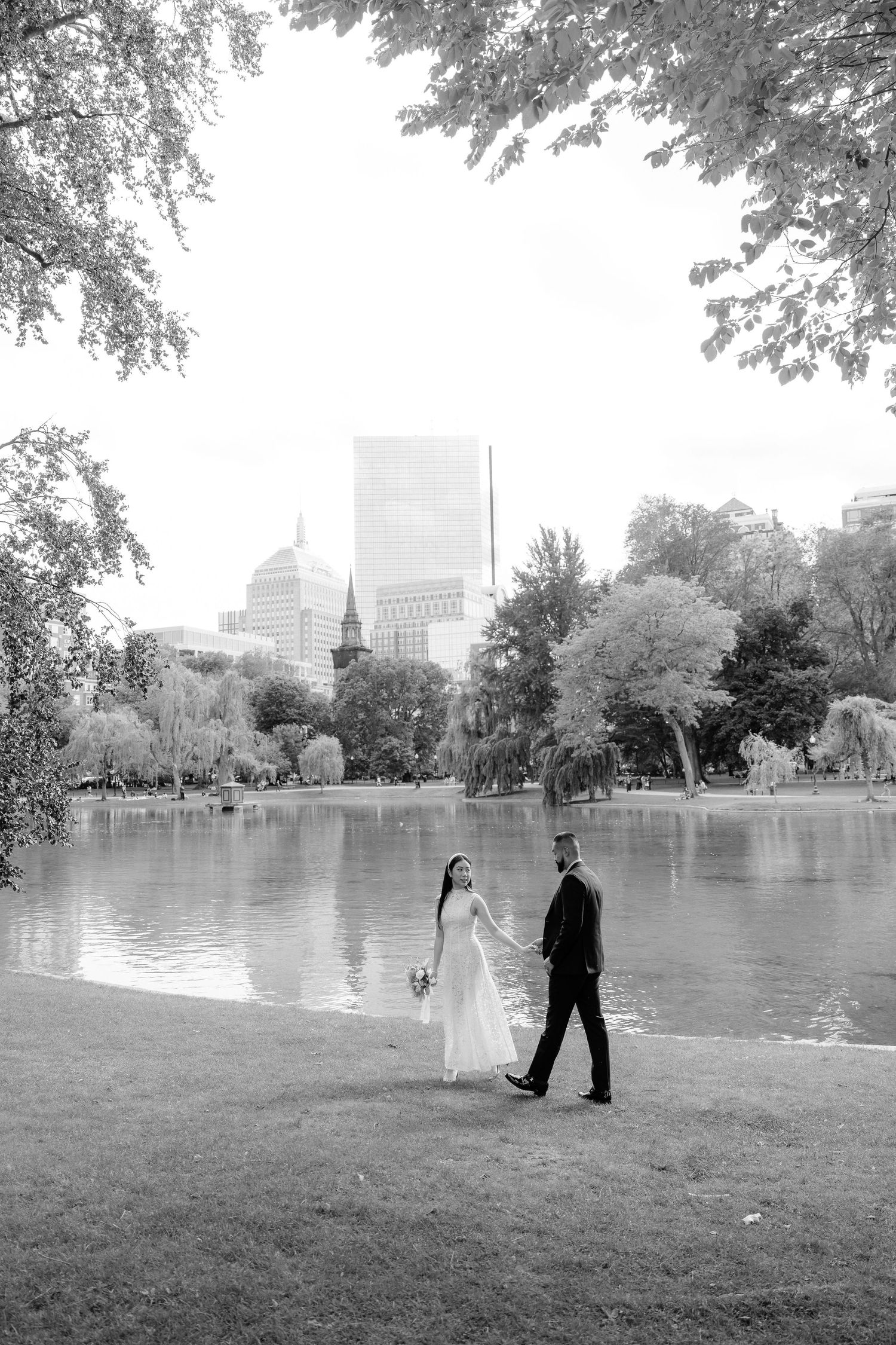 Black and white photo of a couple walking by a pond with city skyline in background.