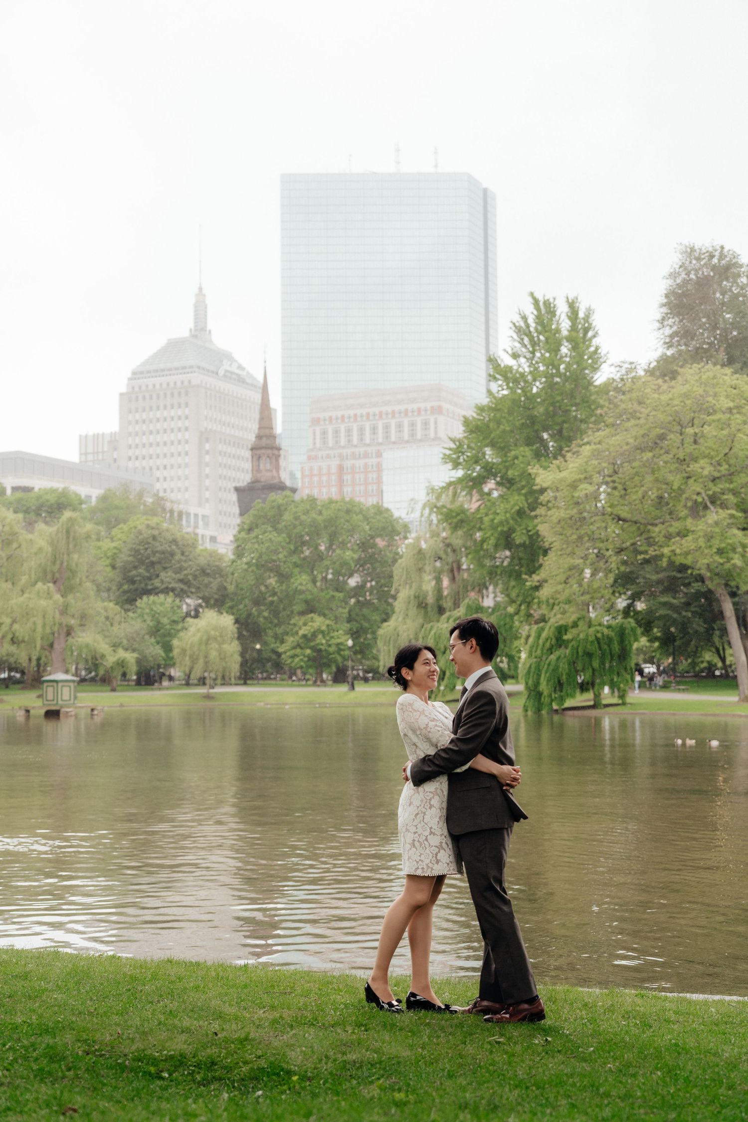 A romantic scene unfolds on grass by a lake with Boston's skyline visible in the background.