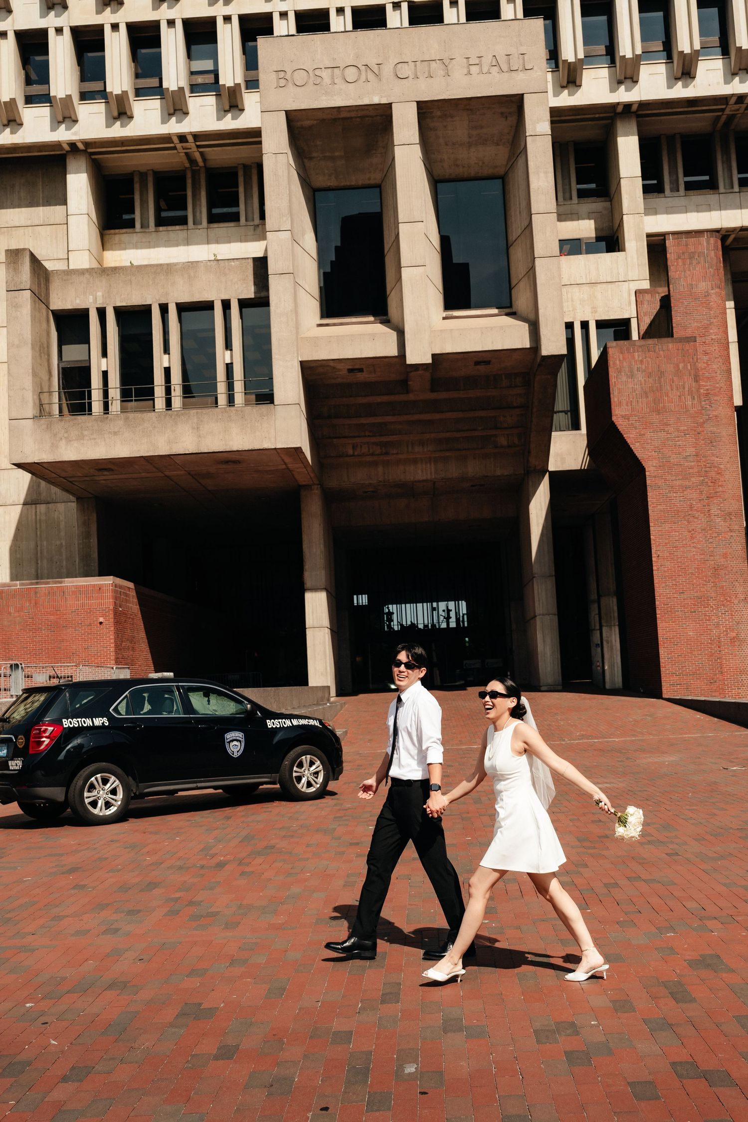 A couple walks across the brick plaza in front of Boston City Hall on a sunny day.
