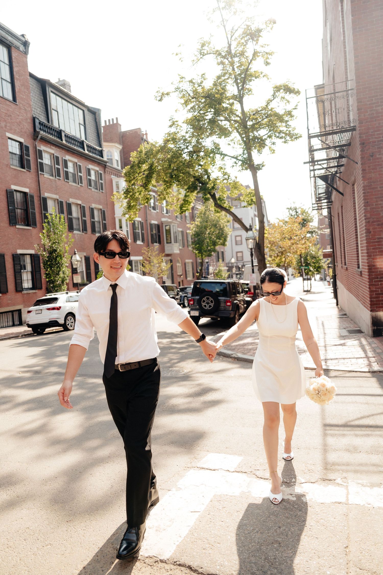 A couple in white and black attire holds hands while crossing a sunny city street.
