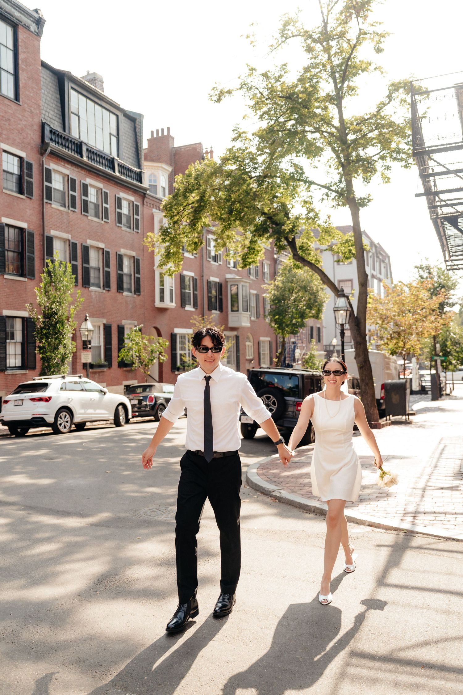 A couple holds hands while walking down a sunny Boston street lined with brownstone buildings.