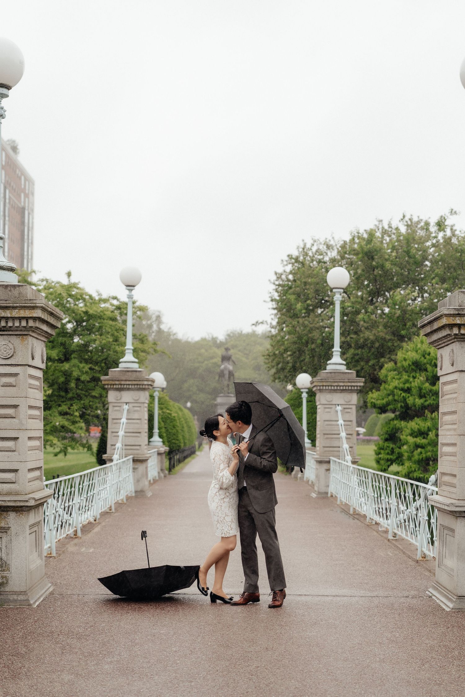 A couple shares an intimate moment on a bridge with white ornate posts in a park setting.
