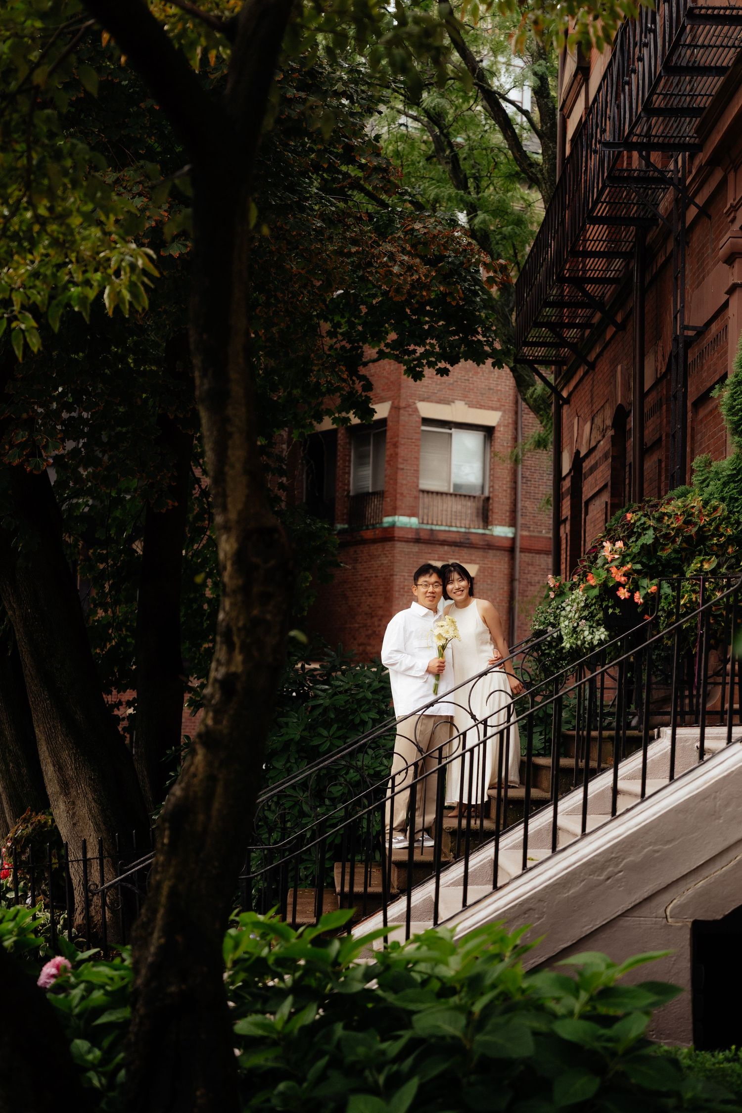 A couple stands on brick brownstone steps surrounded by lush greenery in an urban setting.
