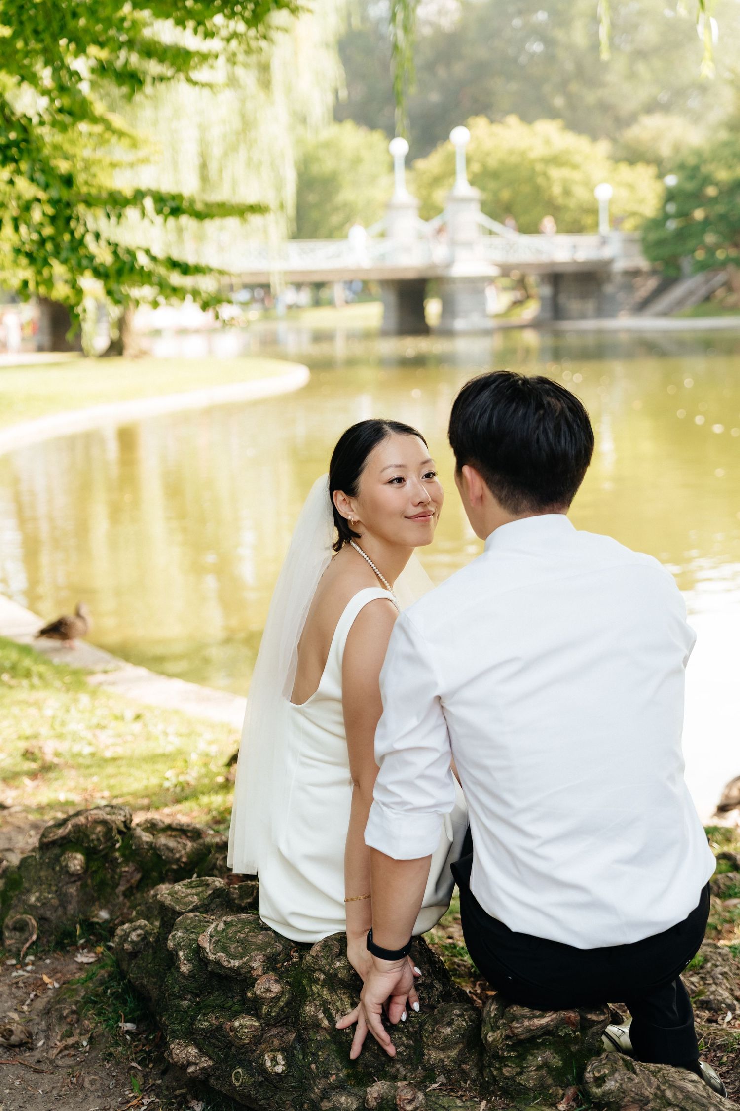 A couple sits by a serene pond with an ornate bridge visible in the background.