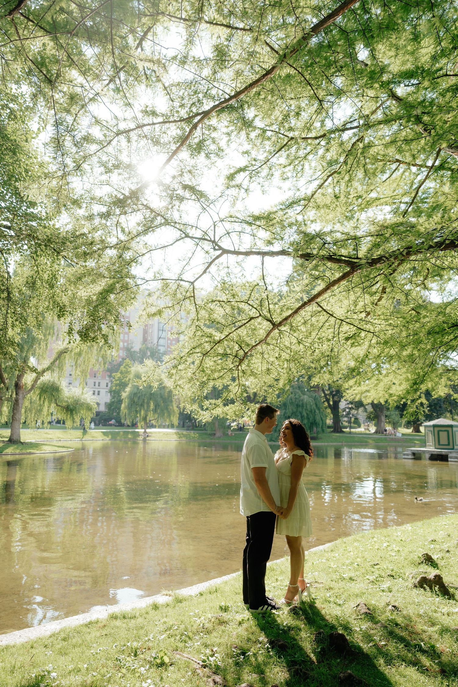 A romantic scene unfolds by a tranquil pond with sunlight filtering through spring trees.