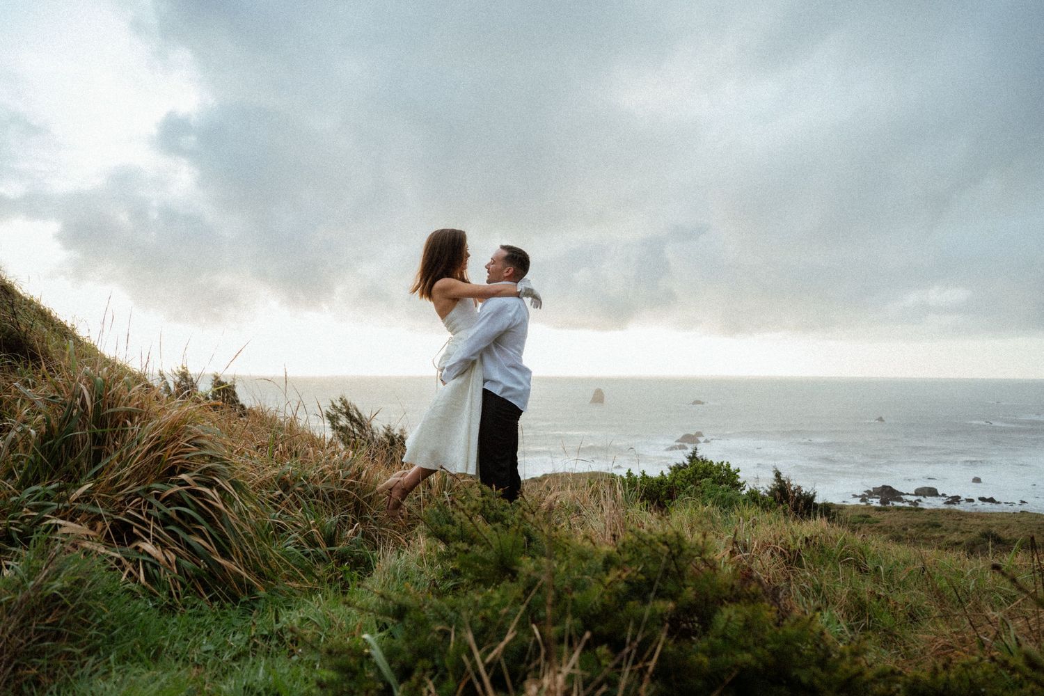A couple embraces on a windswept coastal trail with ocean views.