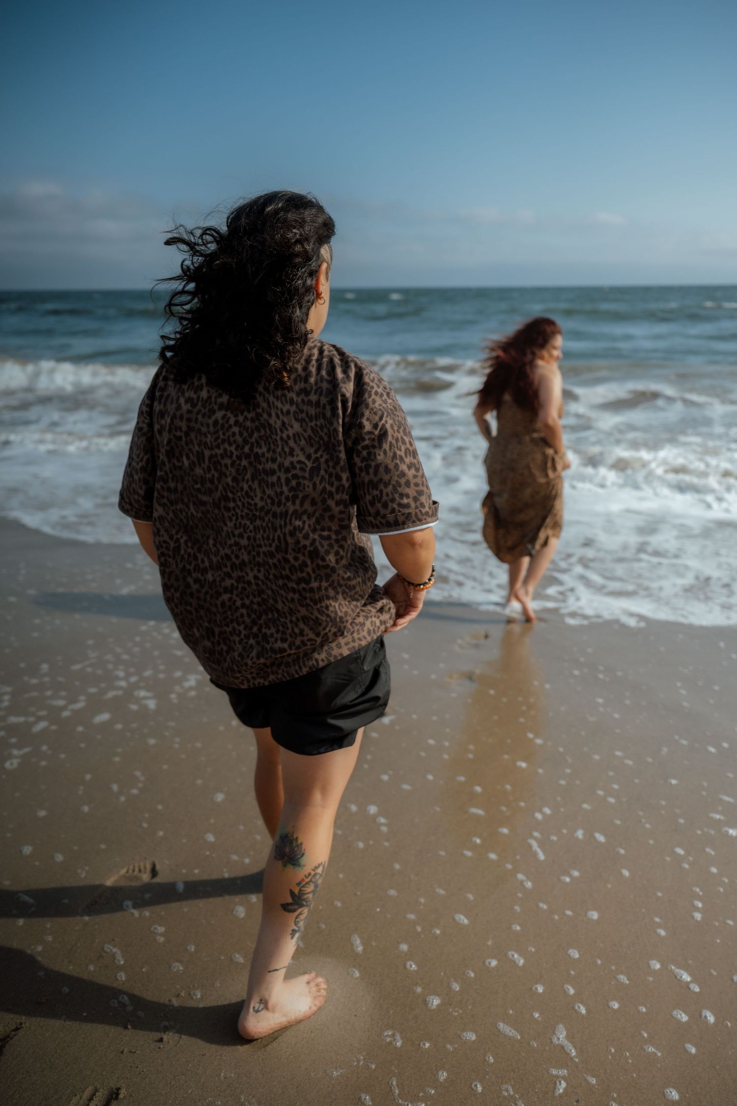 Two people in animal print clothing walking along a sunny beach with waves crashing.