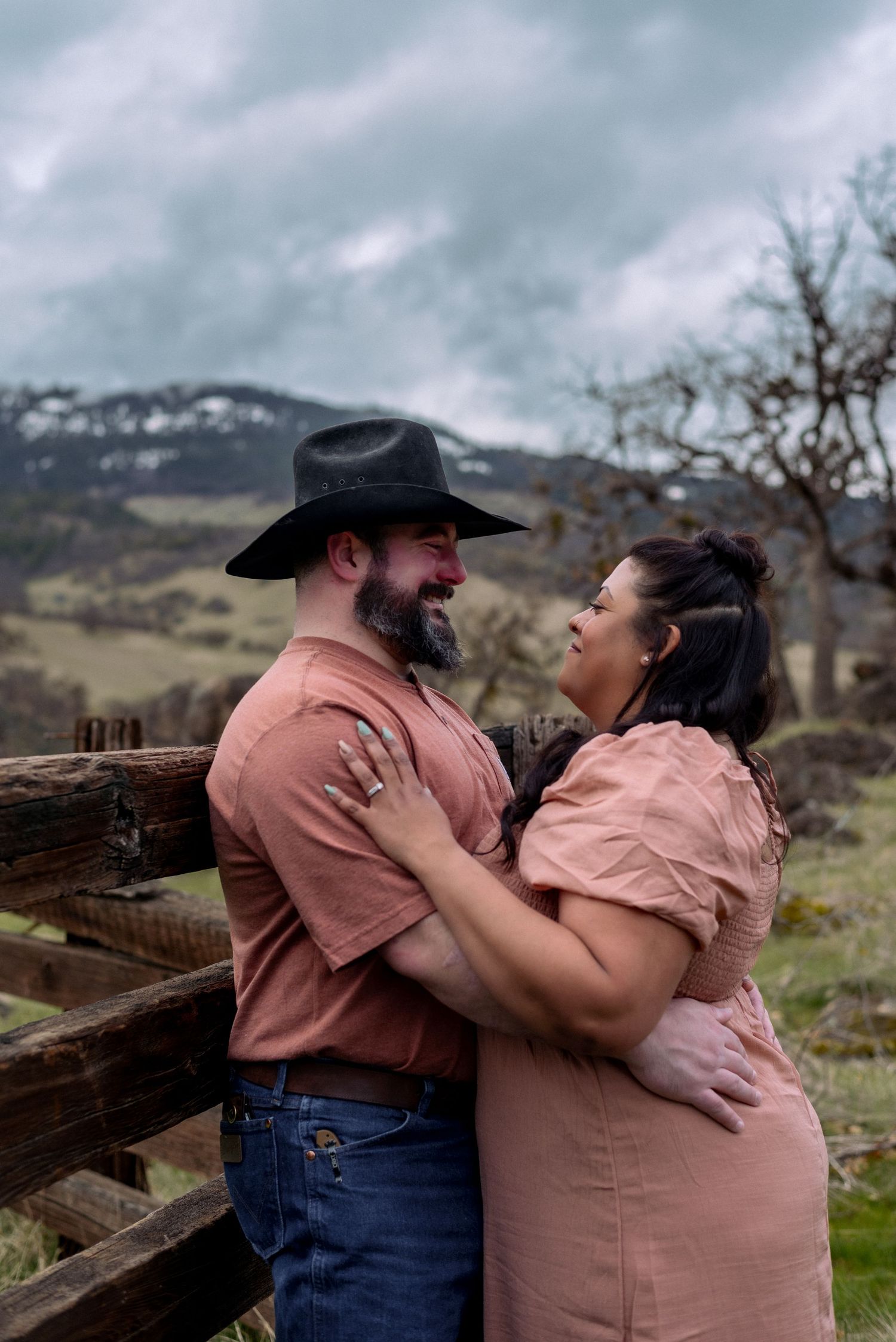 A couple embraces lovingly by a wooden fence in a rural mountain setting.