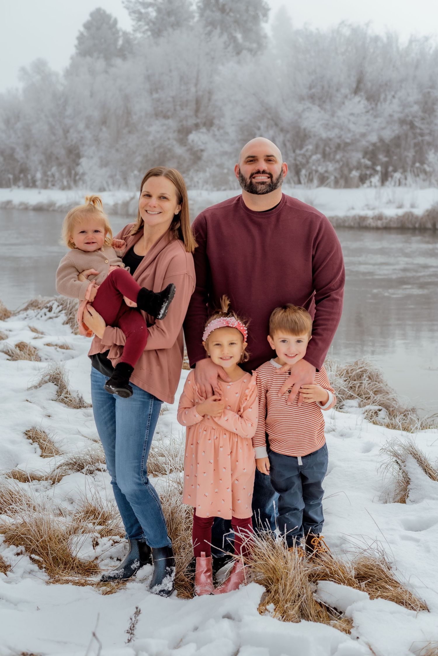 Winter family portrait by frozen lake with coordinated pink and burgundy outfits.